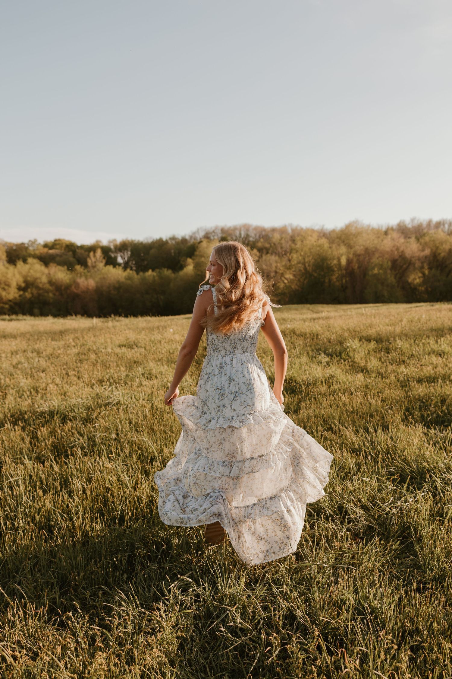 Woman in floral tiered dress twirling in a sunlit green field with trees in the background.