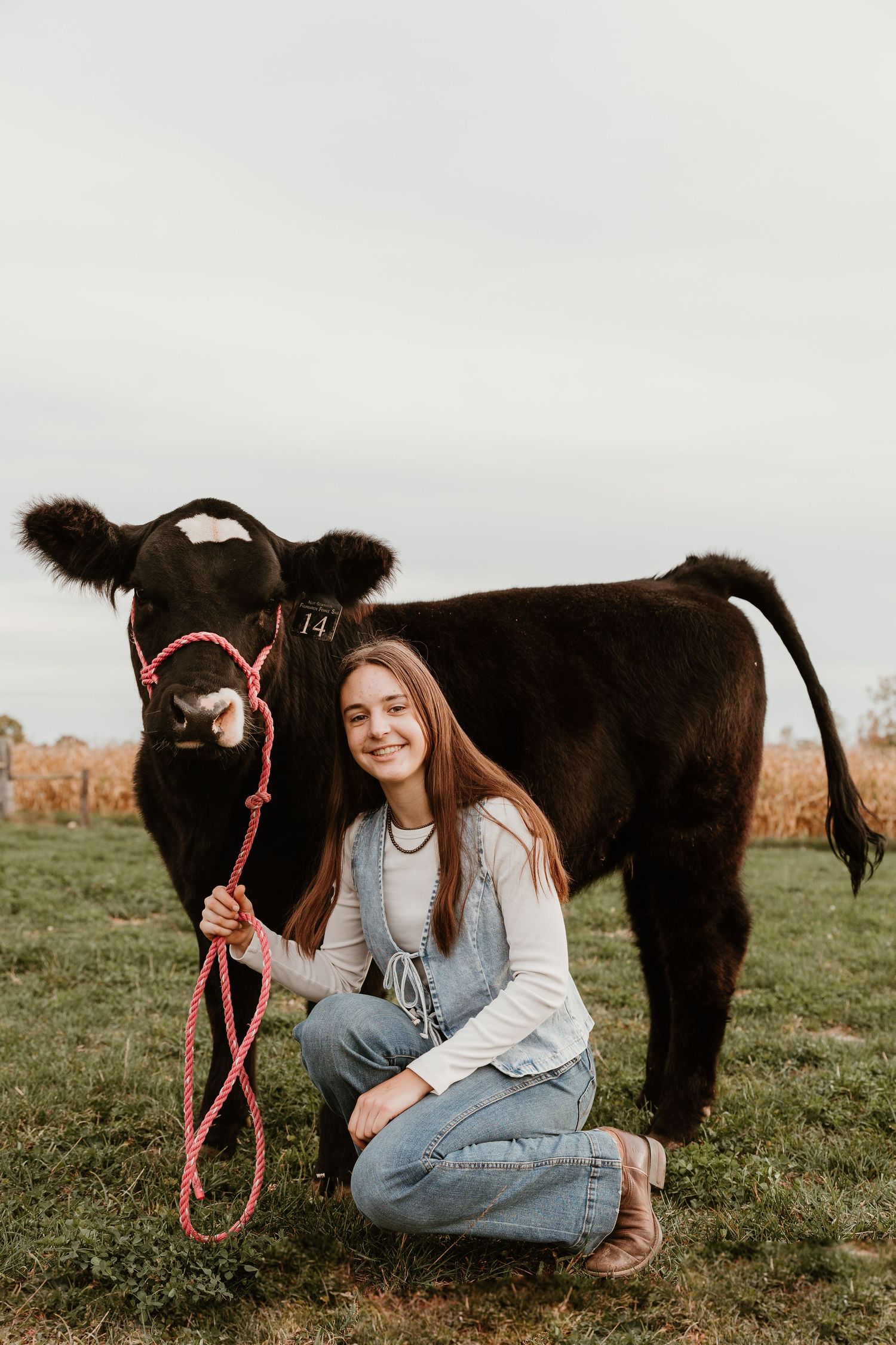 Young woman kneeling beside a black calf on green grass, holding a pink rope halter, smiling outdoors.