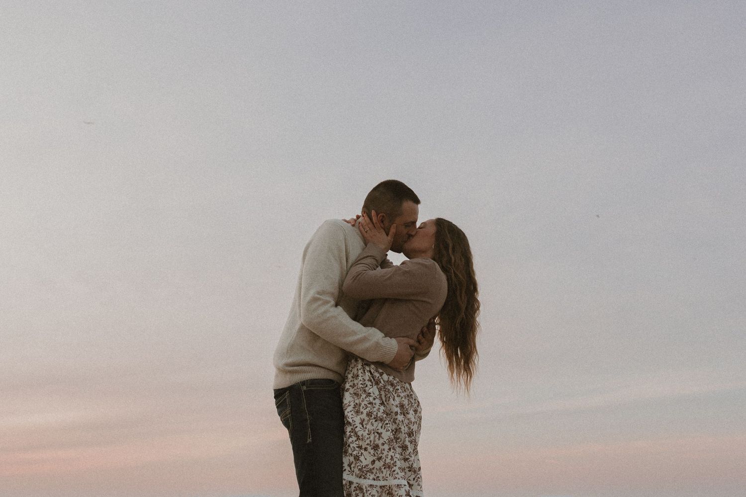 A couple shares a romantic kiss outdoors against a soft pastel sky at dusk, dressed in casual warm tones.