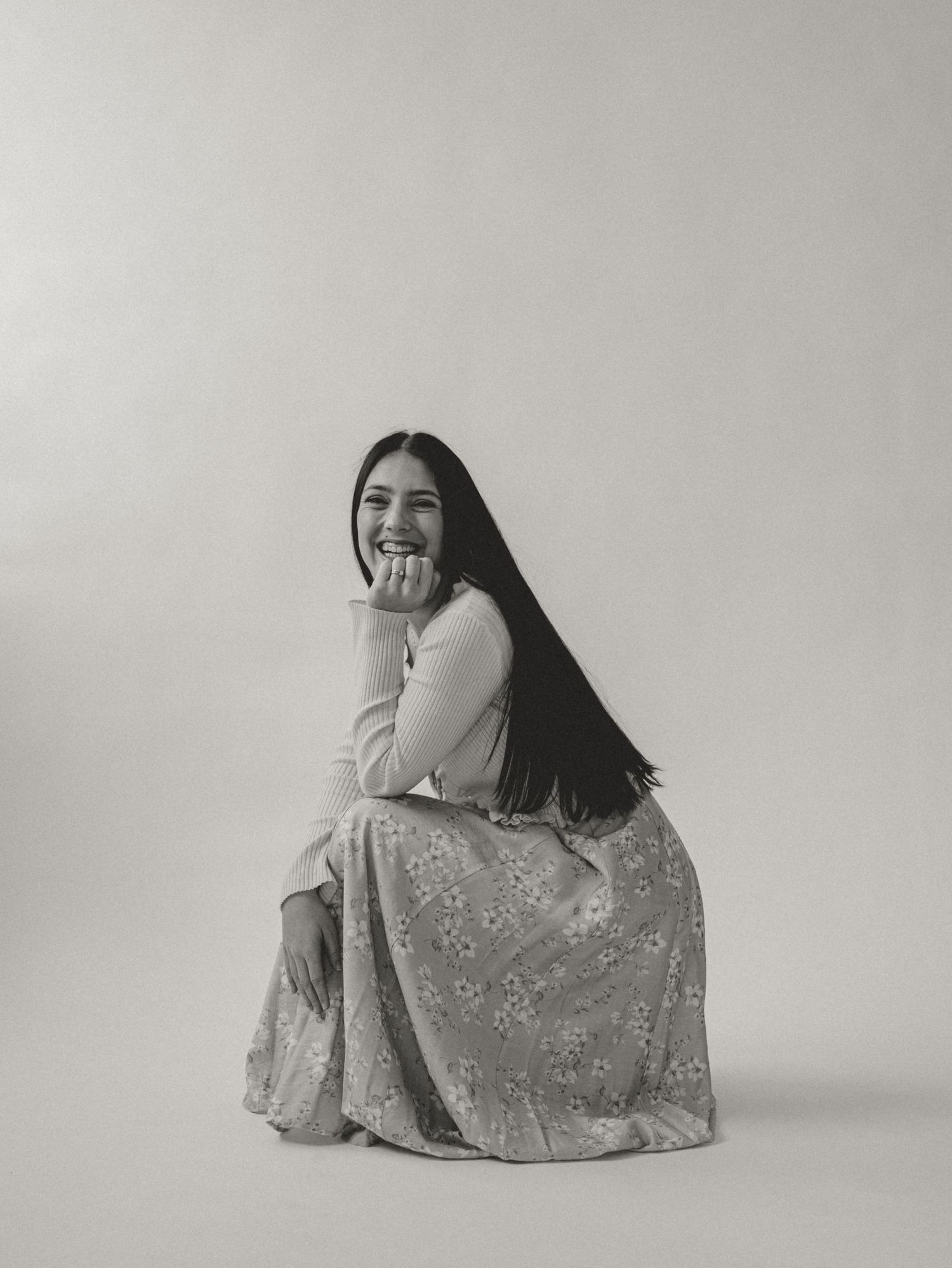 Young woman with long dark hair sits smiling, wearing floral skirt and white top, in black and white portrait.