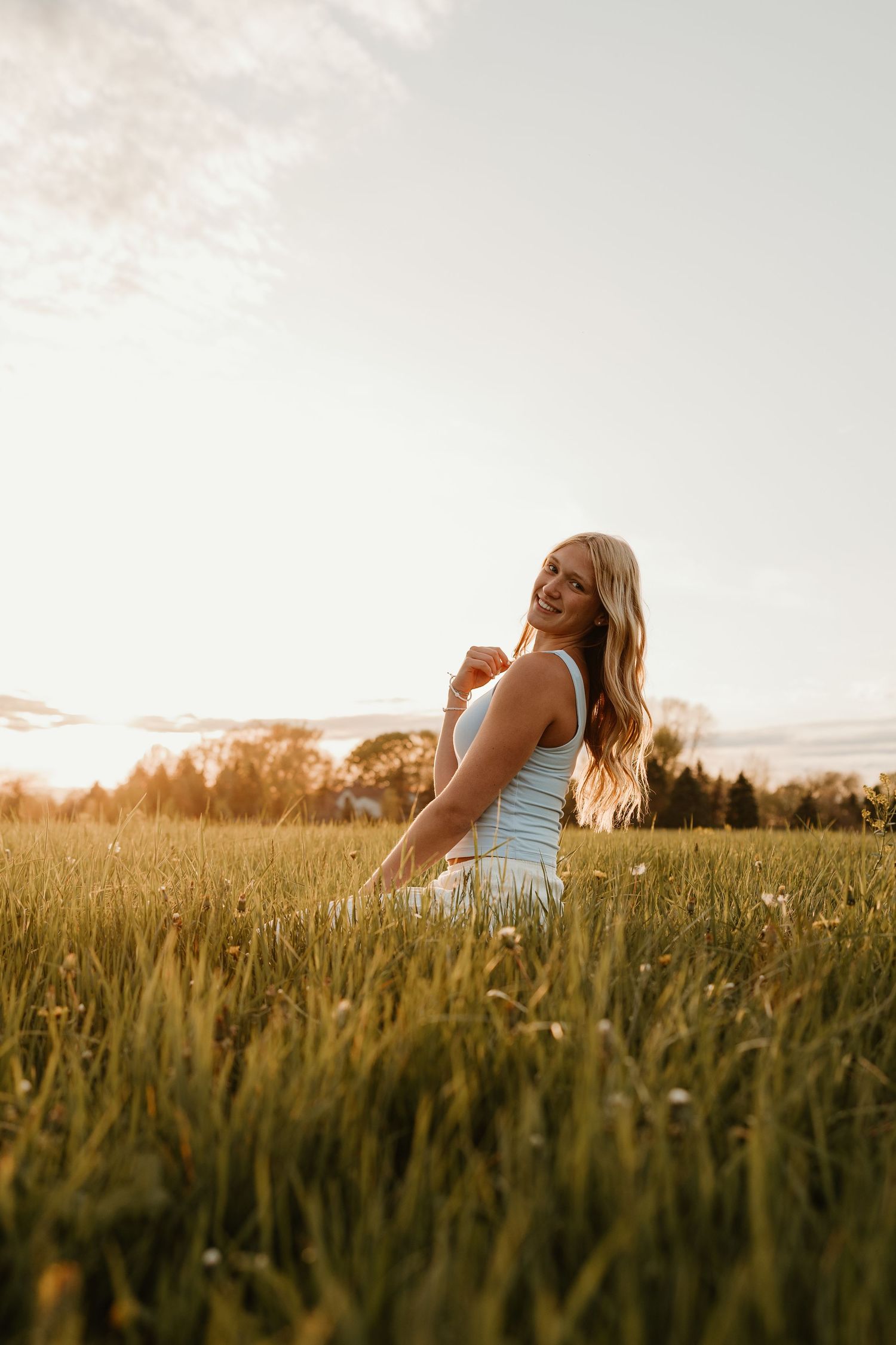 Smiling blonde woman in white tank top sitting in a sunlit green field during golden hour sunset.