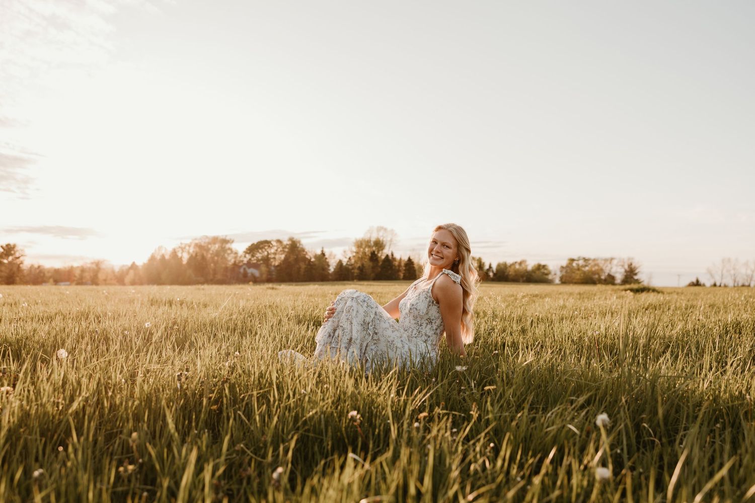 A young woman in a floral dress sits in a golden sunlit field at sunset with trees in the background.
