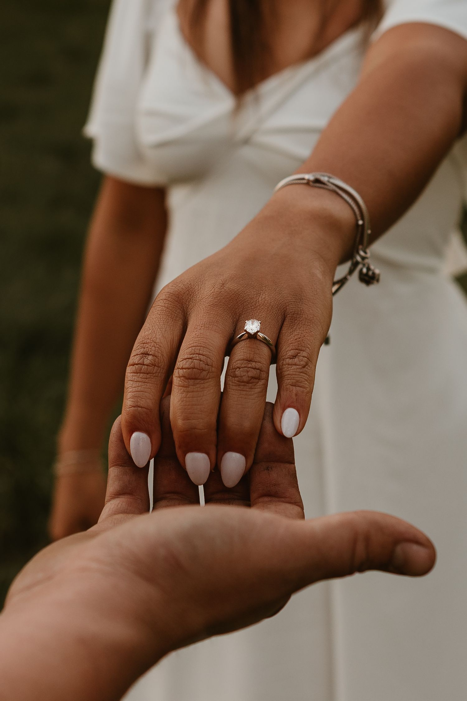 Close-up of a woman in white dress showing off her diamond engagement ring while holding hands with her partner outdoors.