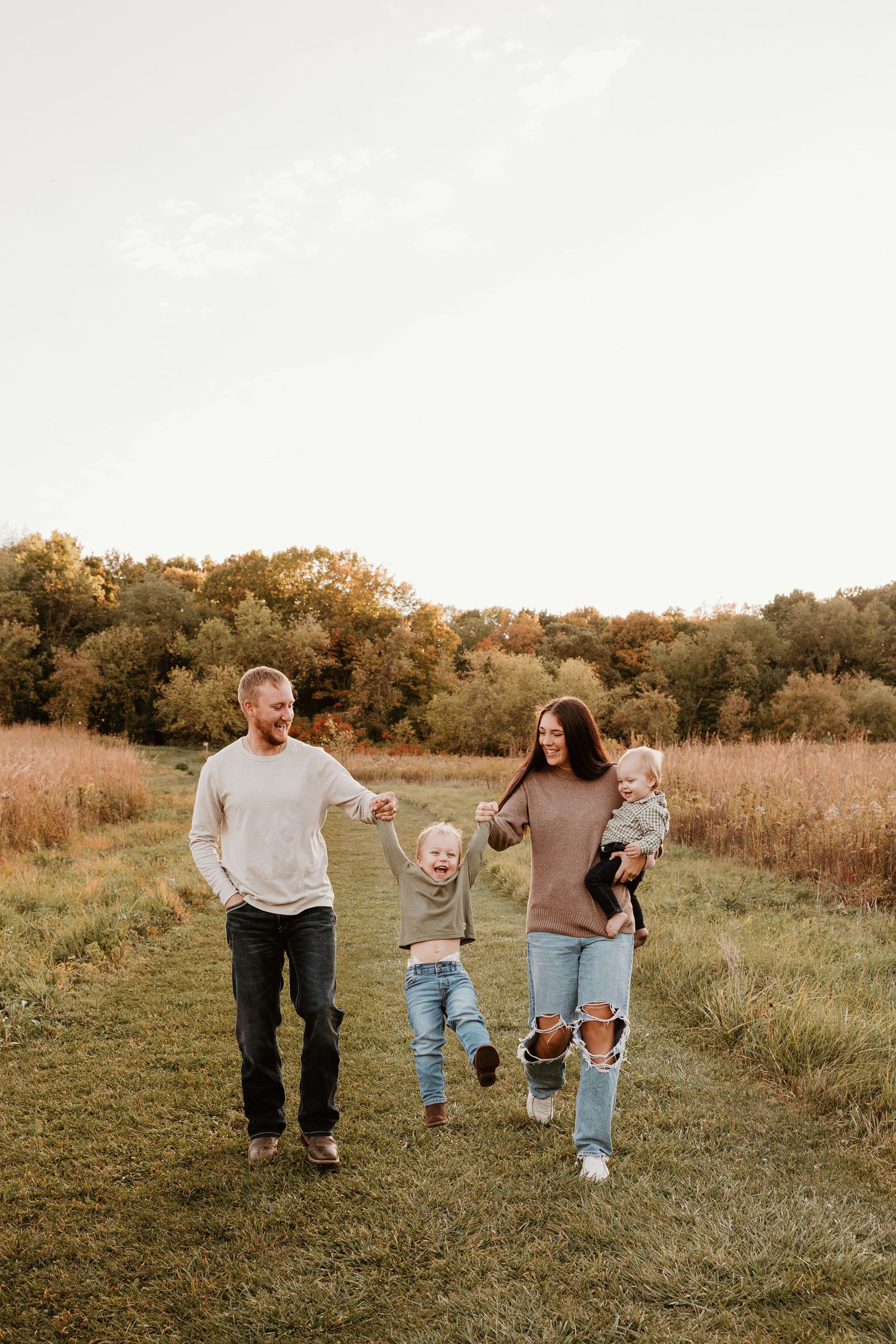Family of four walking in a golden field at sunset, parents swinging toddler between them while holding baby.