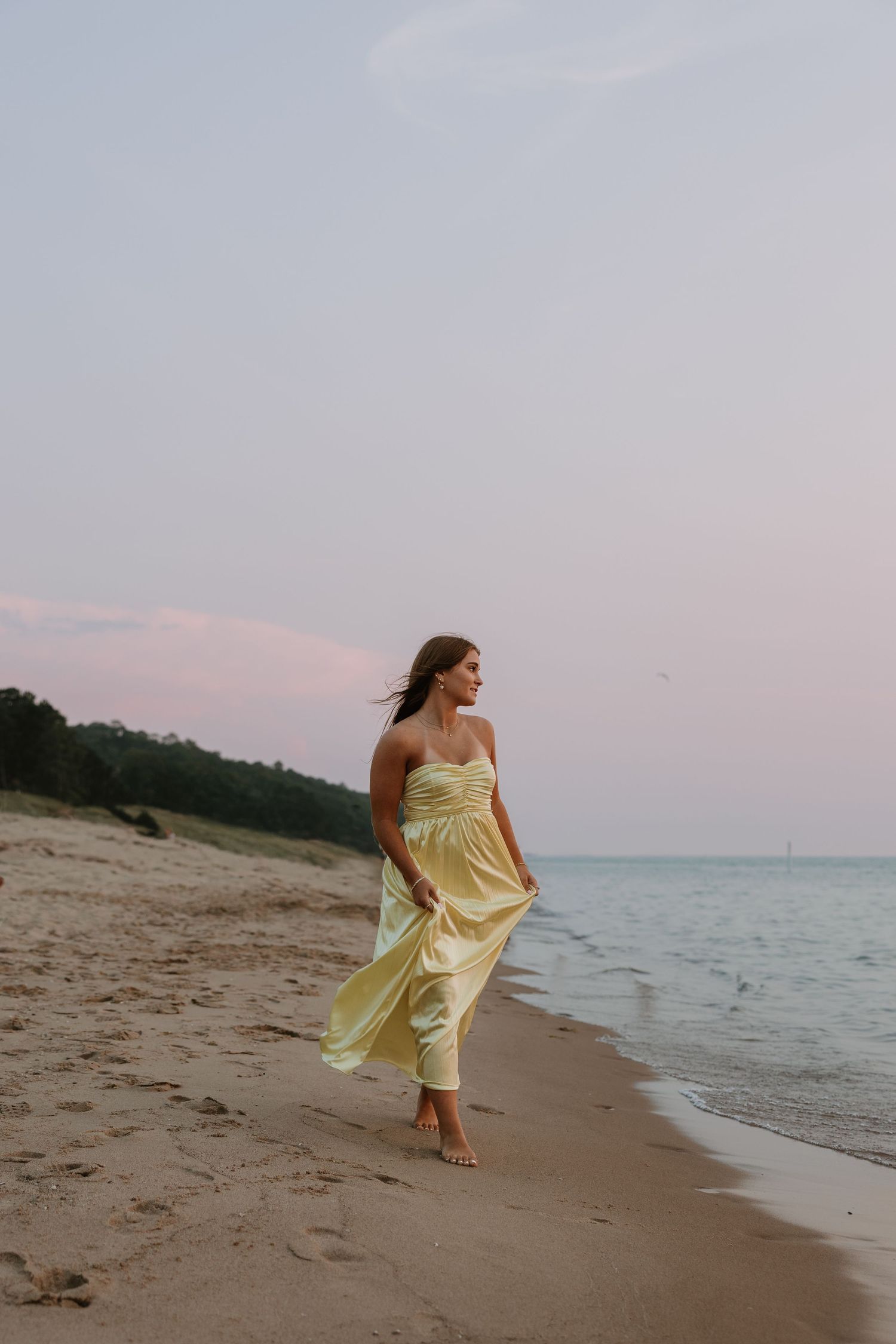 Woman in yellow strapless dress walking barefoot on sandy beach at sunset with calm ocean waters.