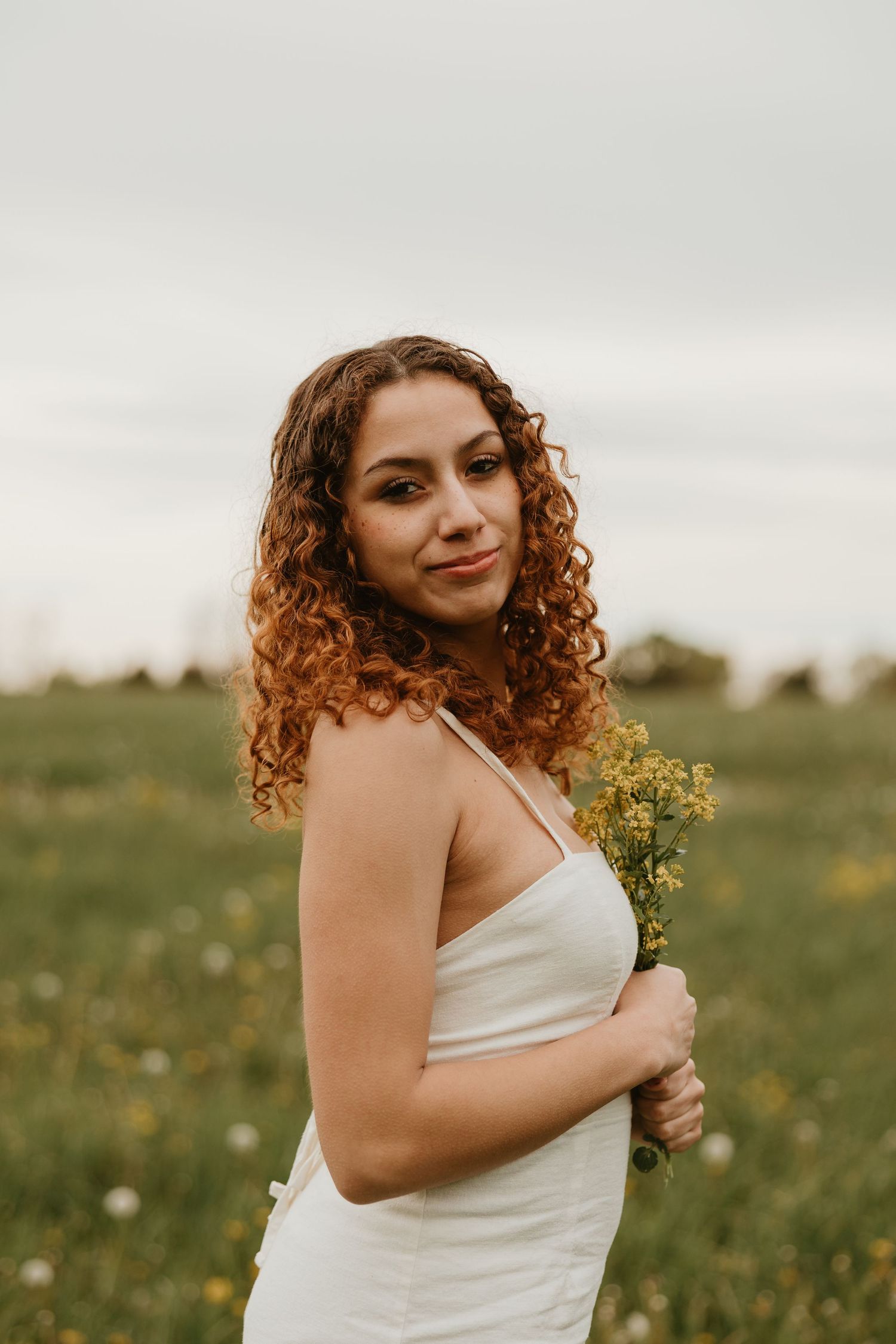 Young woman with curly auburn hair in white dress holding wildflowers in a green field.