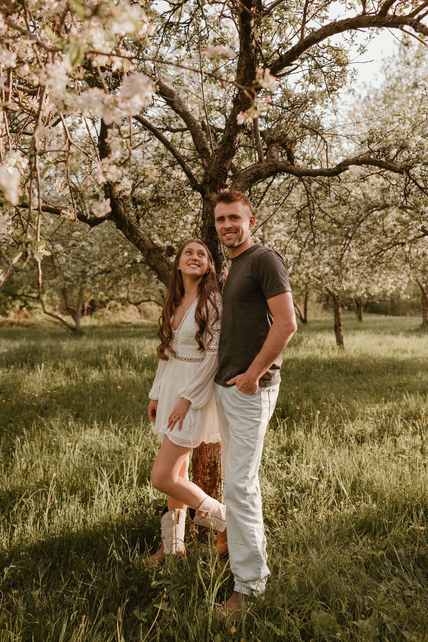 A smiling couple poses together under blooming spring trees in a lush green garden, woman in white dress, man in gray tee.