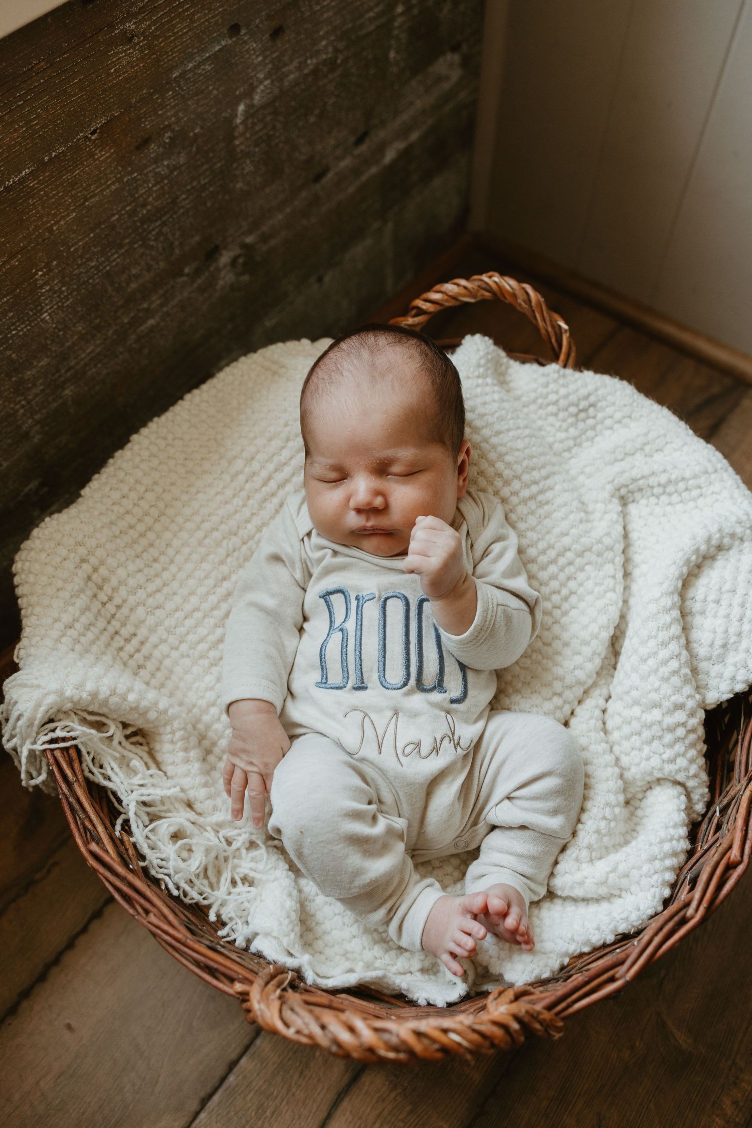 Sleeping newborn baby in personalized onesie resting in wicker basket with white knit blanket.
