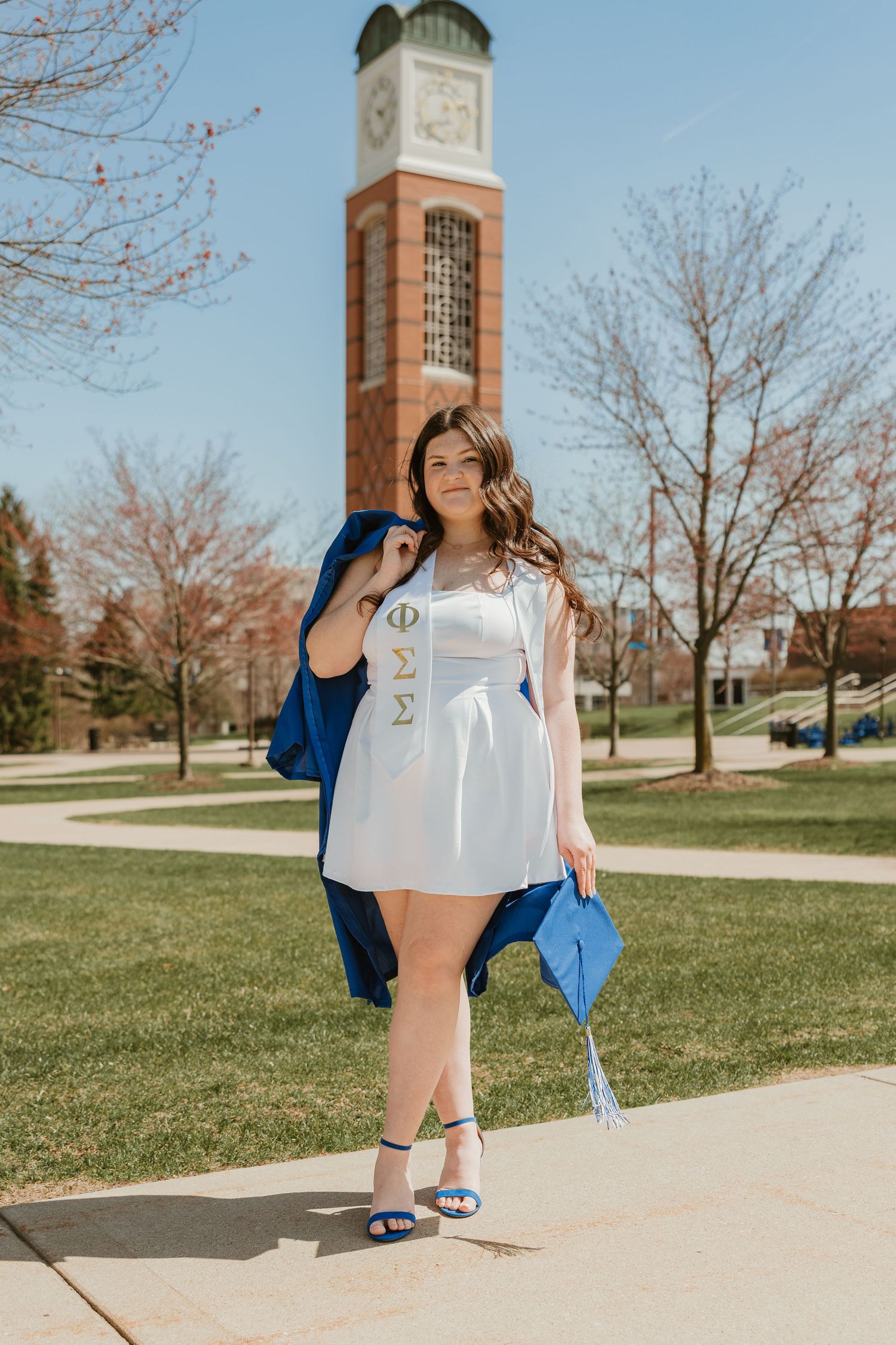College graduate in white dress holds blue cap and gown near brick clock tower on sunny campus.
