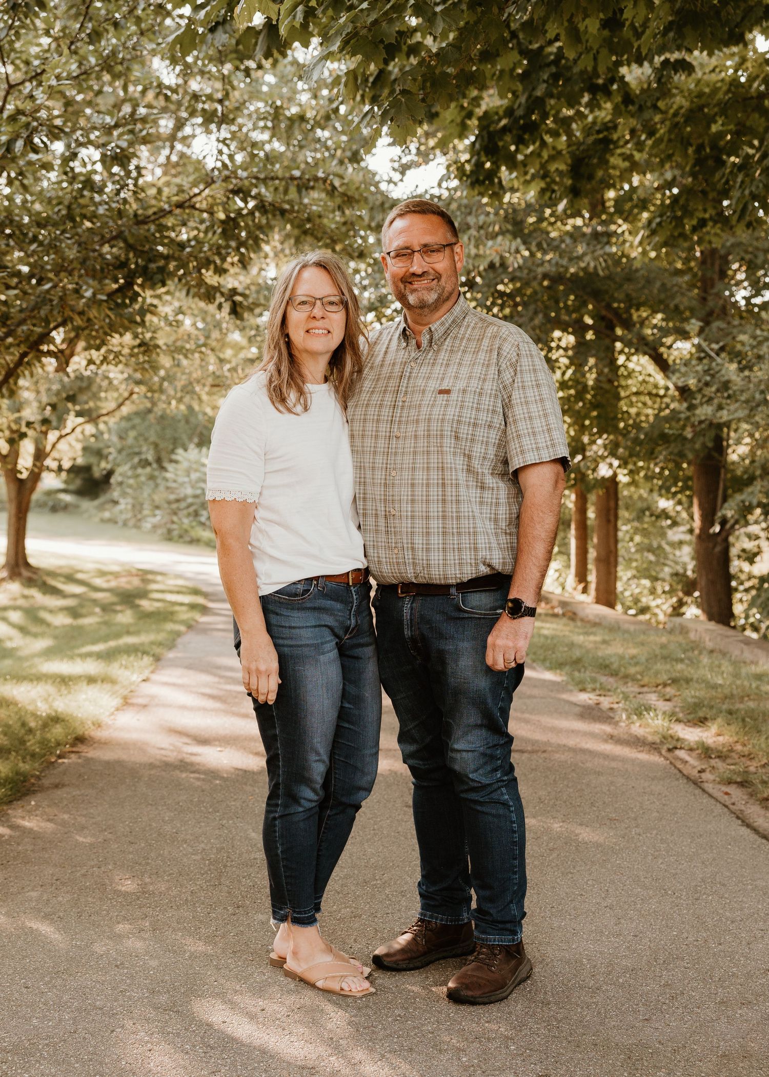 A couple smiles together on a tree-lined path outdoors, woman in white top and jeans, man in plaid shirt and jeans.