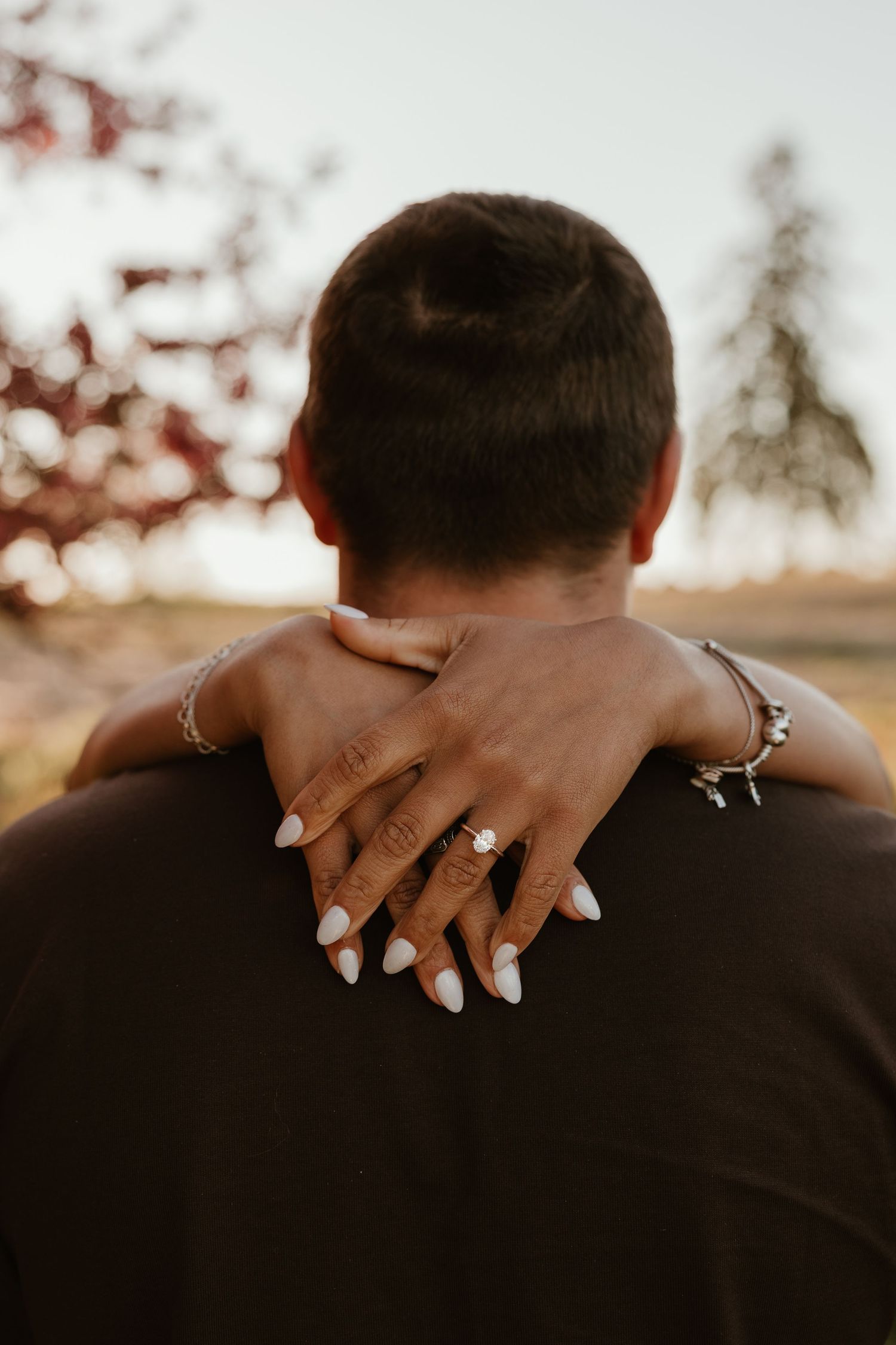 Woman with engagement ring and bracelets embracing man from behind outdoors at golden hour.