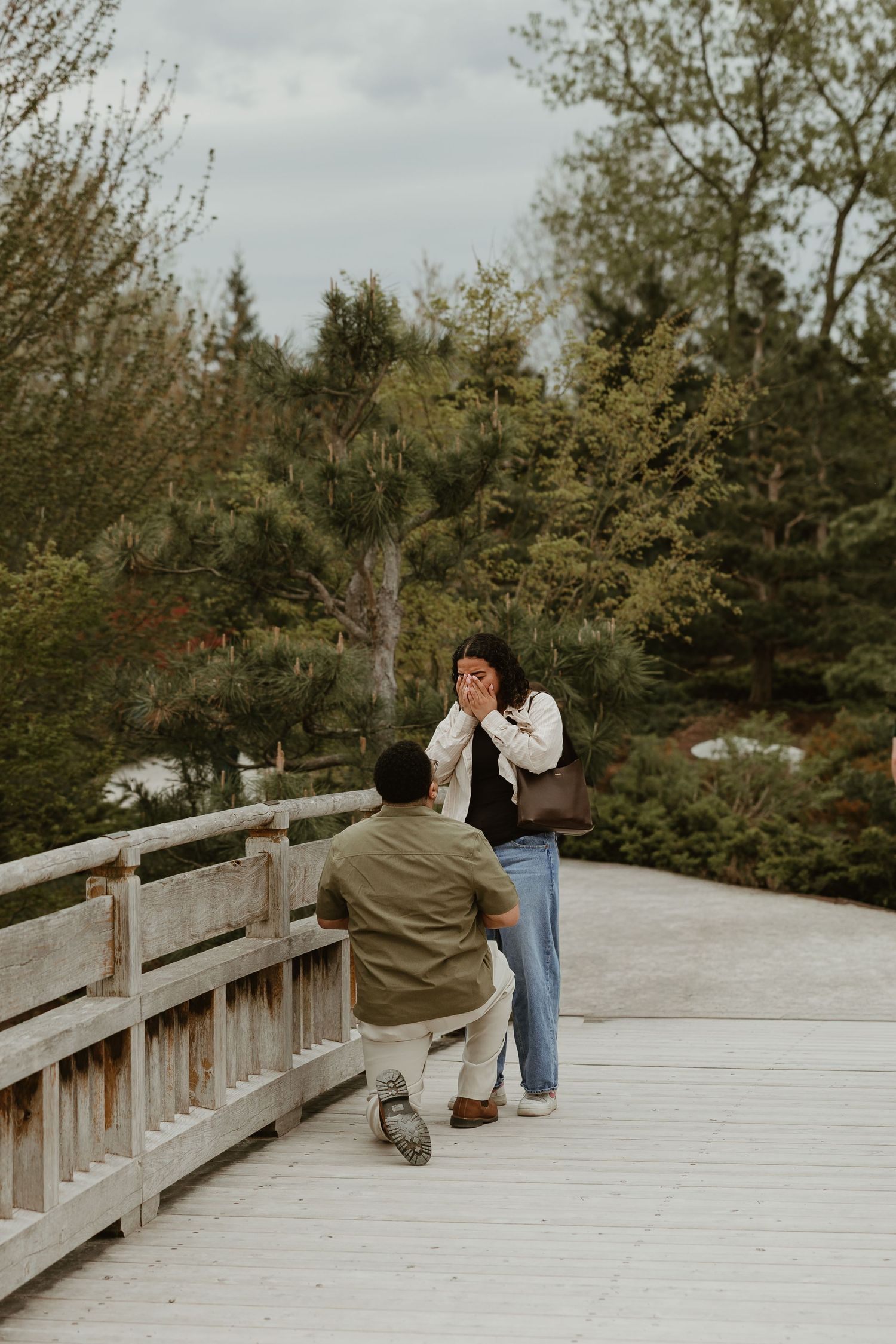 Man kneeling on wooden bridge proposing to surprised woman covering her mouth, surrounded by lush green trees.