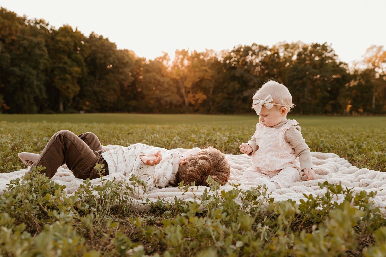 Two young siblings playing on a blanket in a sunlit field at golden hour, surrounded by lush green grass and trees.
