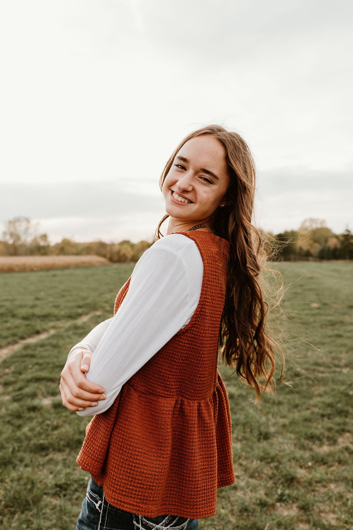 Smiling young woman with long brown hair wearing a rust orange vest over white blouse standing in a green field.