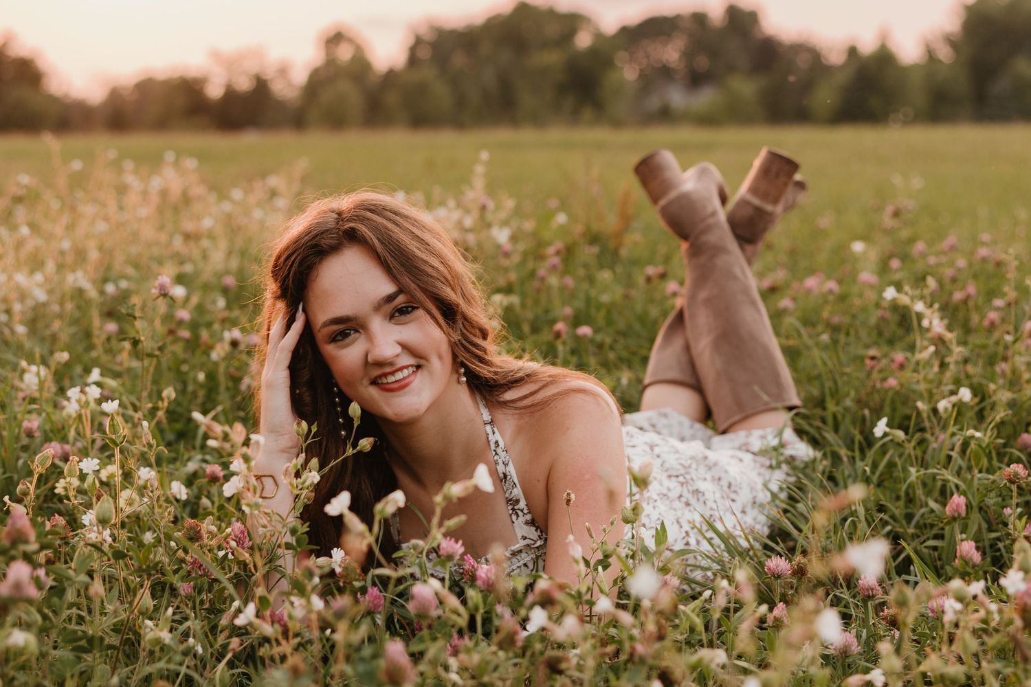 A smiling young woman with auburn hair lies in a wildflower meadow at golden hour, wearing a white dress and brown boots.