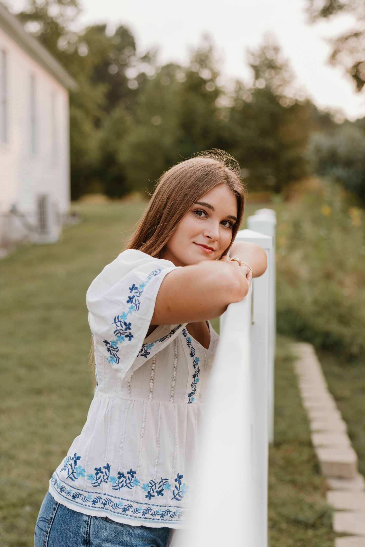Young woman in white embroidered blouse leans on white fence, smiling outdoors in a lush green yard.