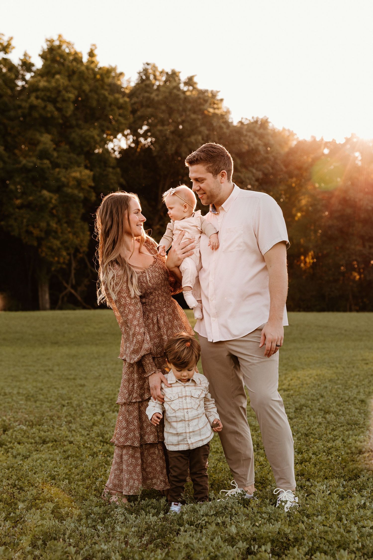 Family of four poses in golden hour sunlight on green field, parents holding baby and toddler, trees in background.