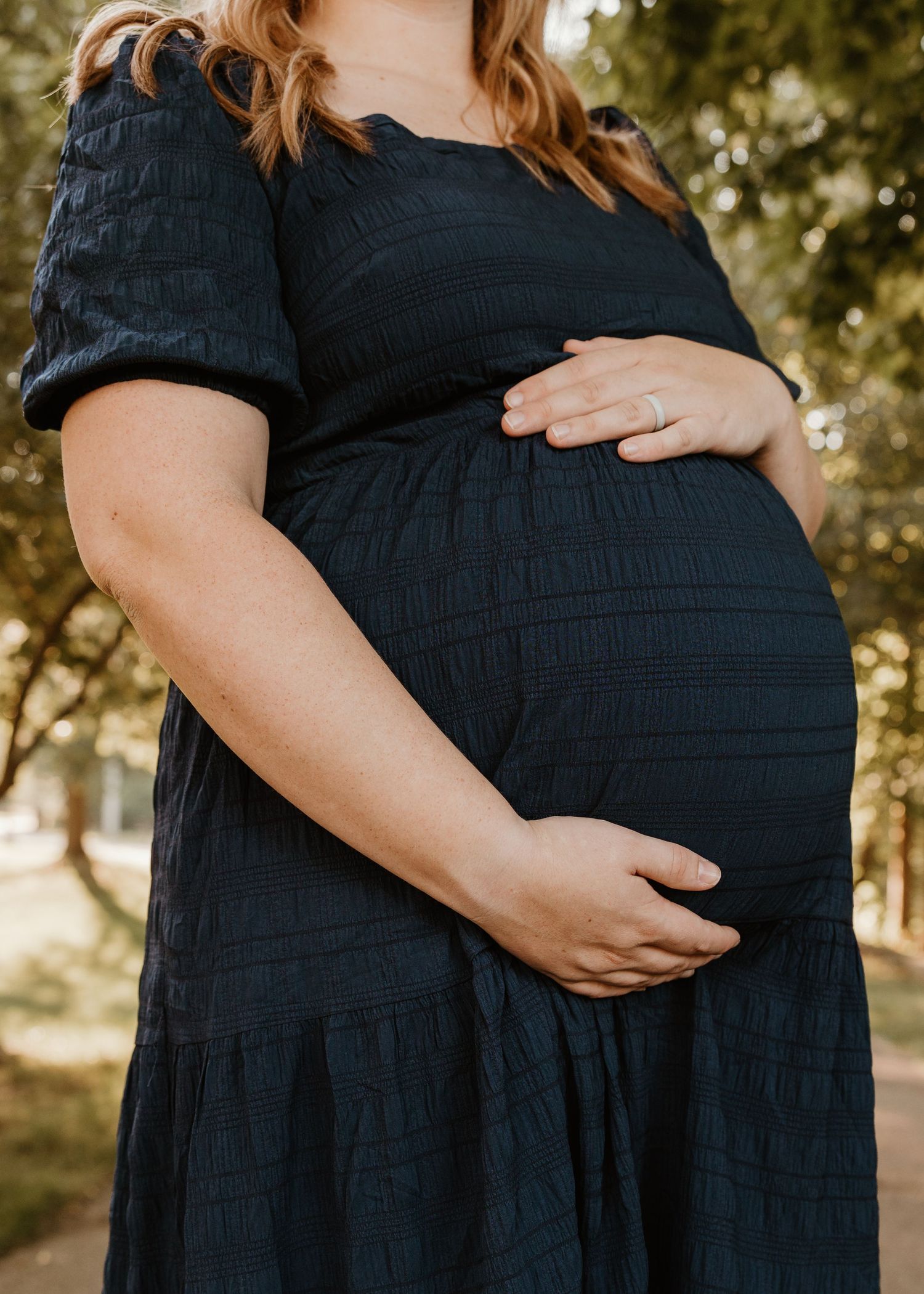 Pregnant woman in black textured dress cradling baby bump outdoors, wearing engagement ring, maternity photo session.
