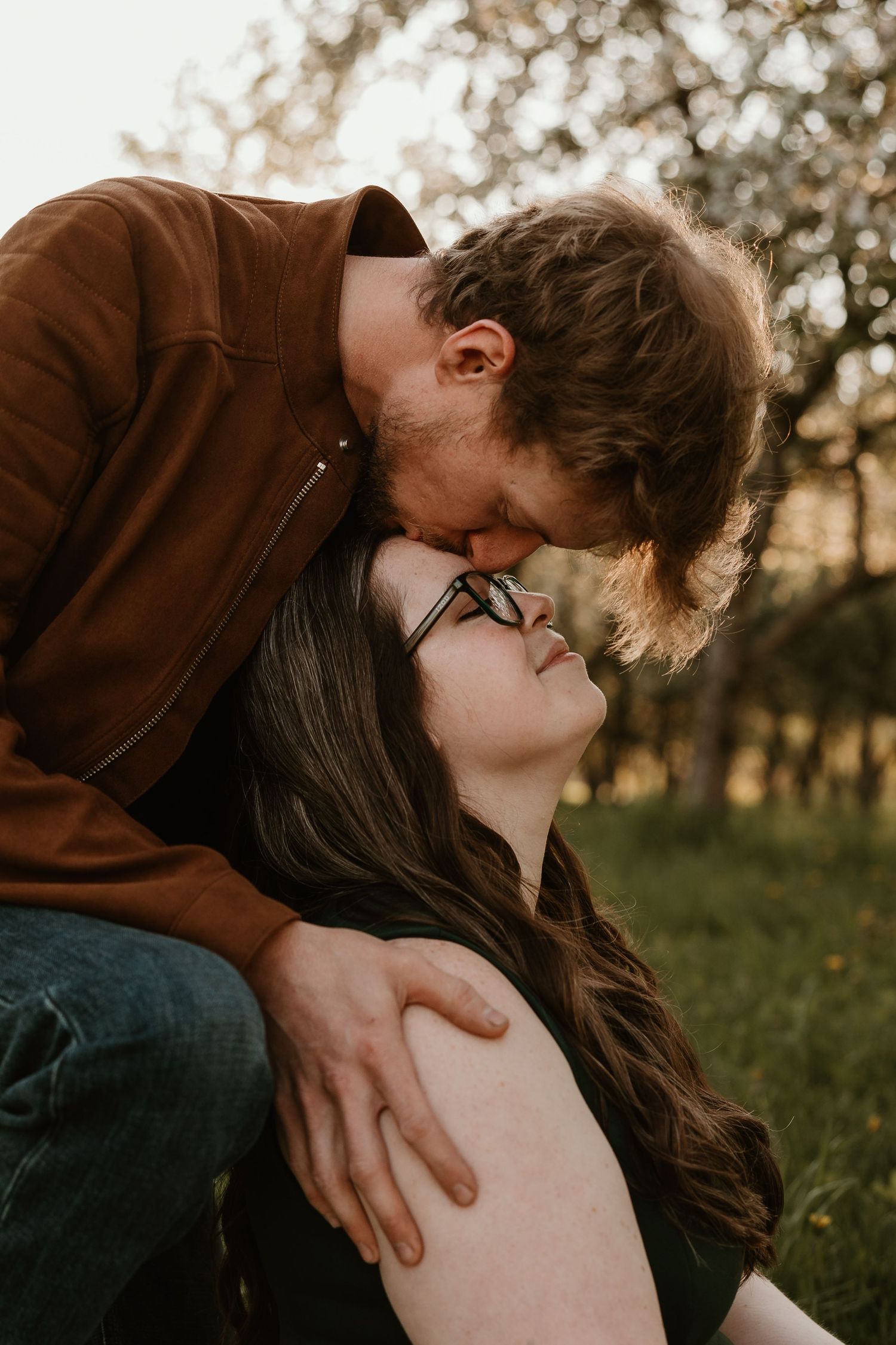 A couple sharing a tender kiss outdoors, woman in glasses leaning back as man in brown jacket embraces her in a green garden.