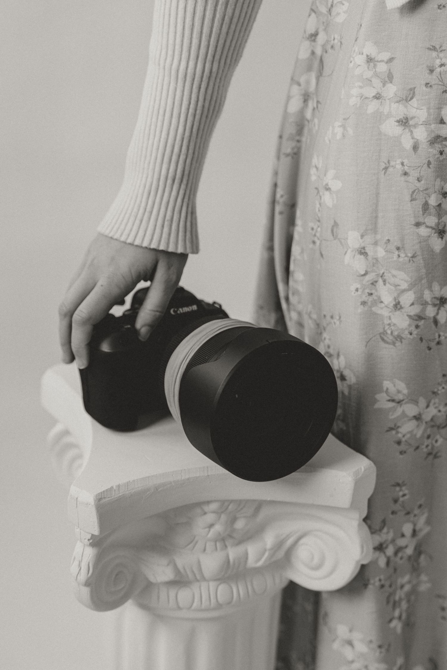 A person holds a Canon DSLR camera with telephoto lens on a white decorative pedestal in a black and white photo.