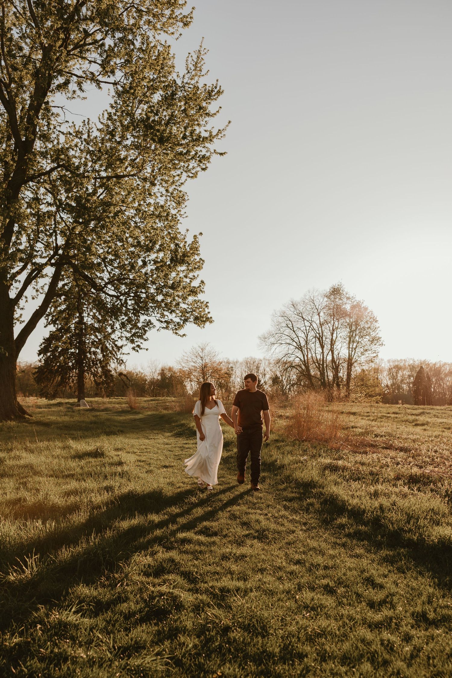 Couple walking hand in hand through a sunlit green field at golden hour with trees in the background.