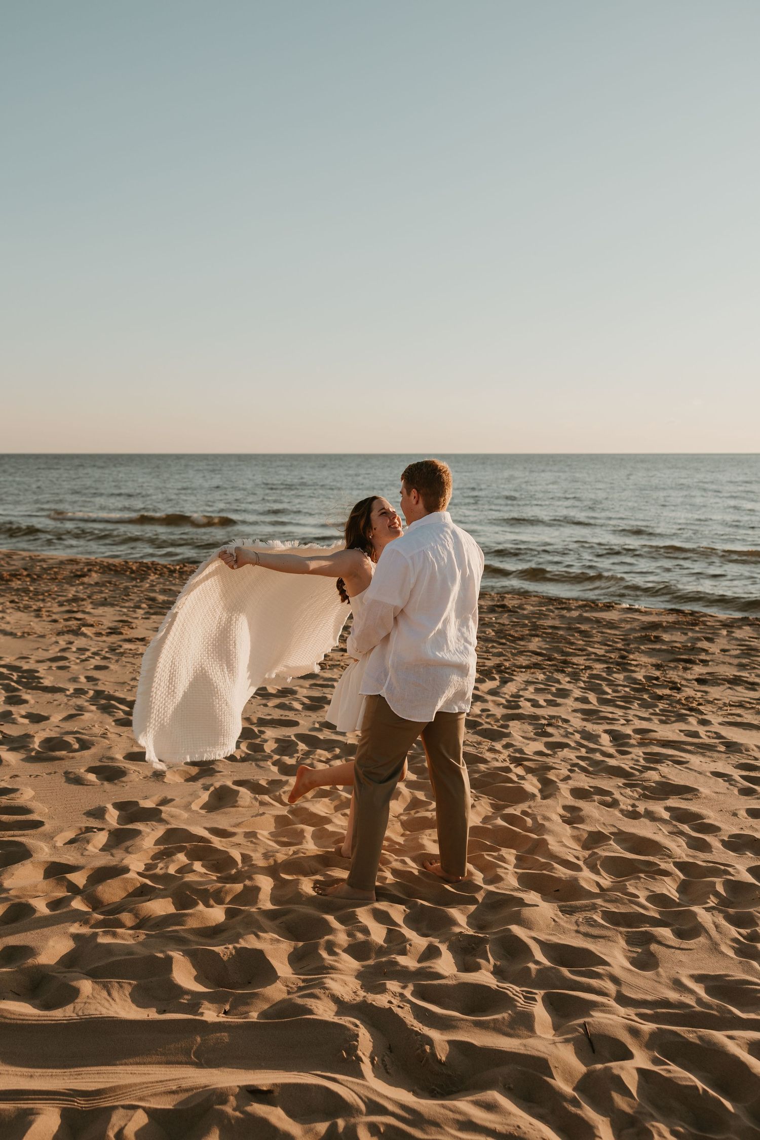 Couple in white outfits embracing on sandy beach at sunset with ocean in background.