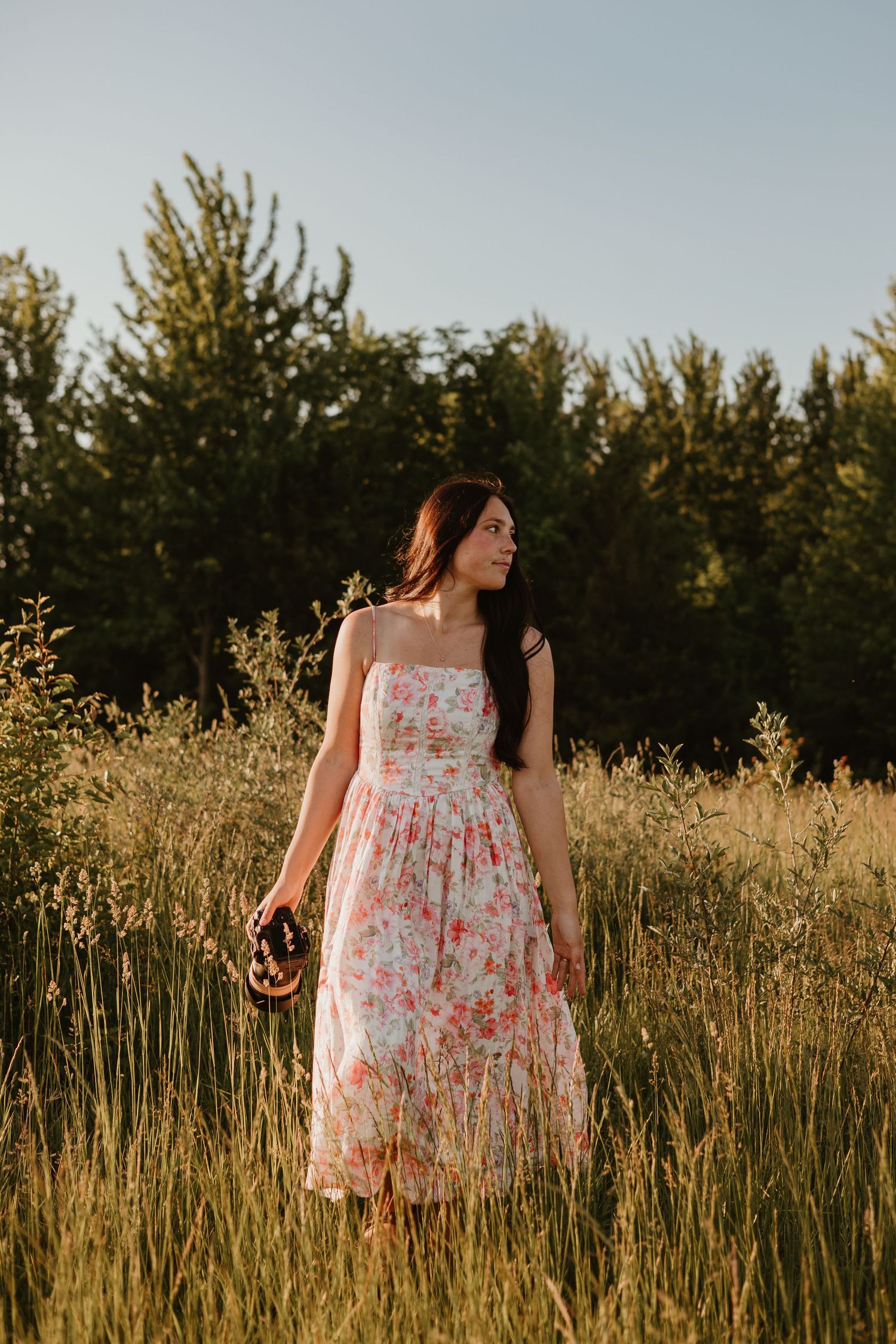 Woman in floral sundress standing in a sunlit meadow with green trees in the background.