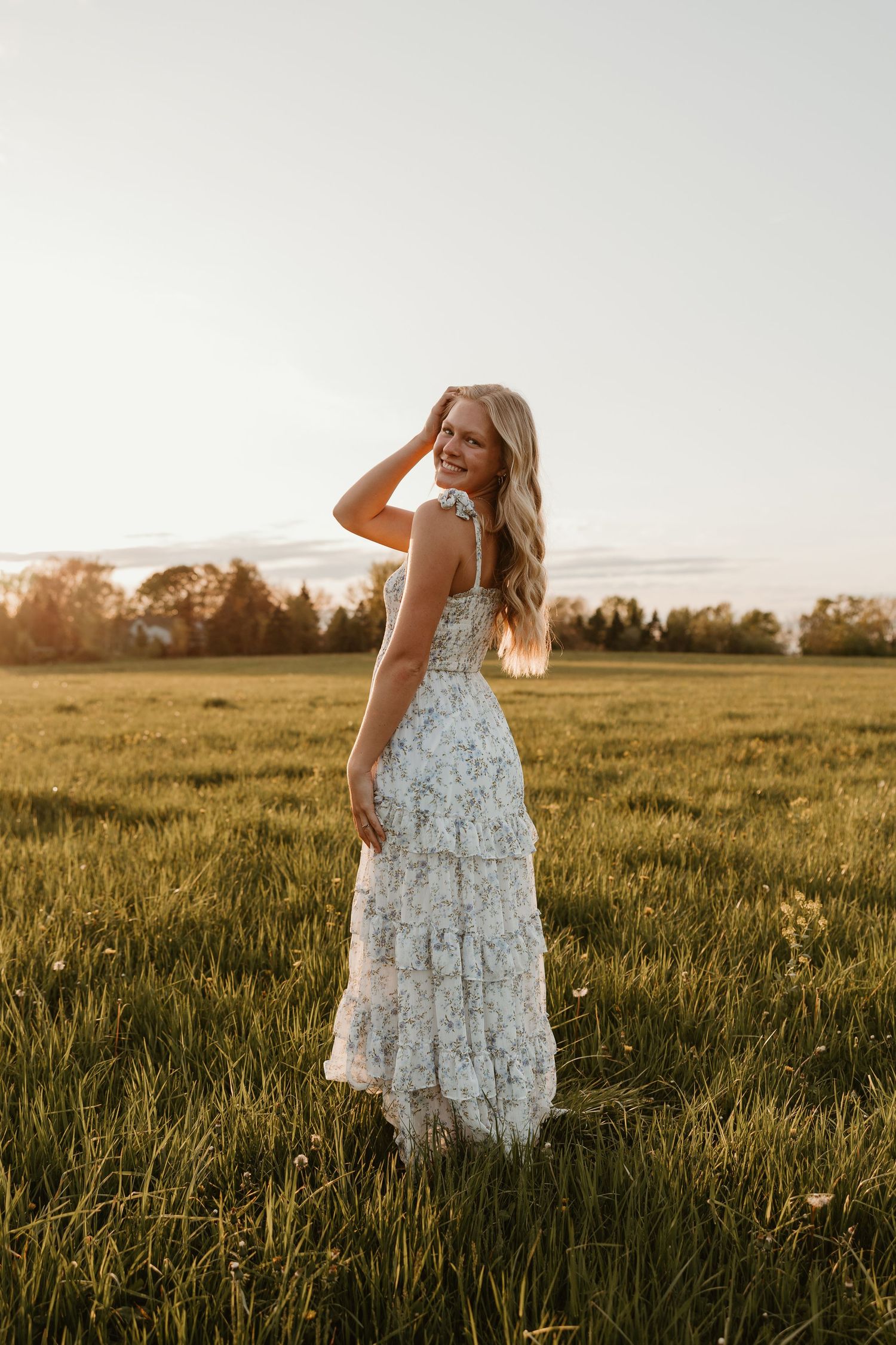 Blonde woman in floral maxi dress standing in green field at golden hour sunset, smiling over shoulder.