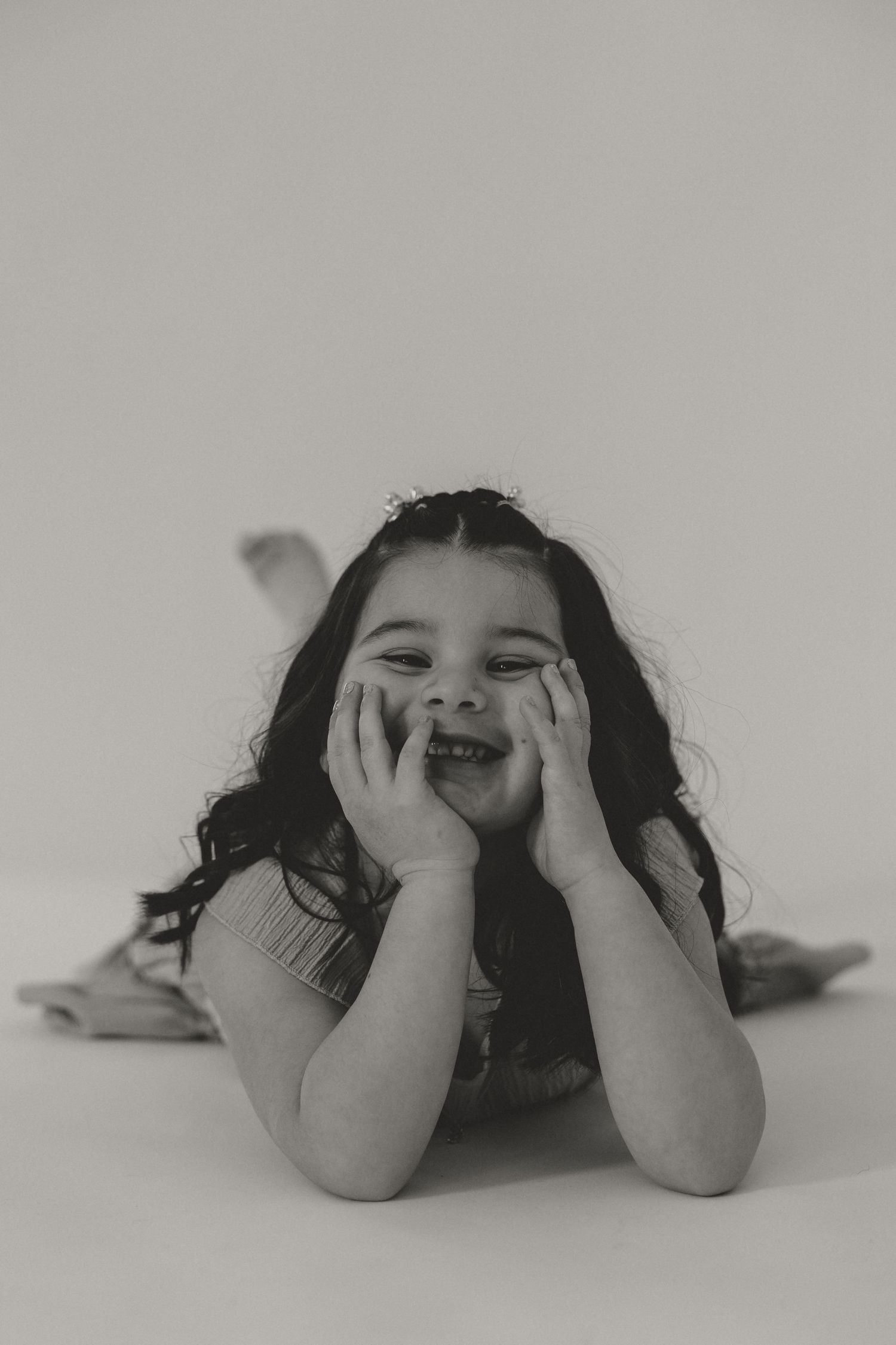 Black and white photo of a cute little girl lying down, resting her face in her hands, smiling playfully.
