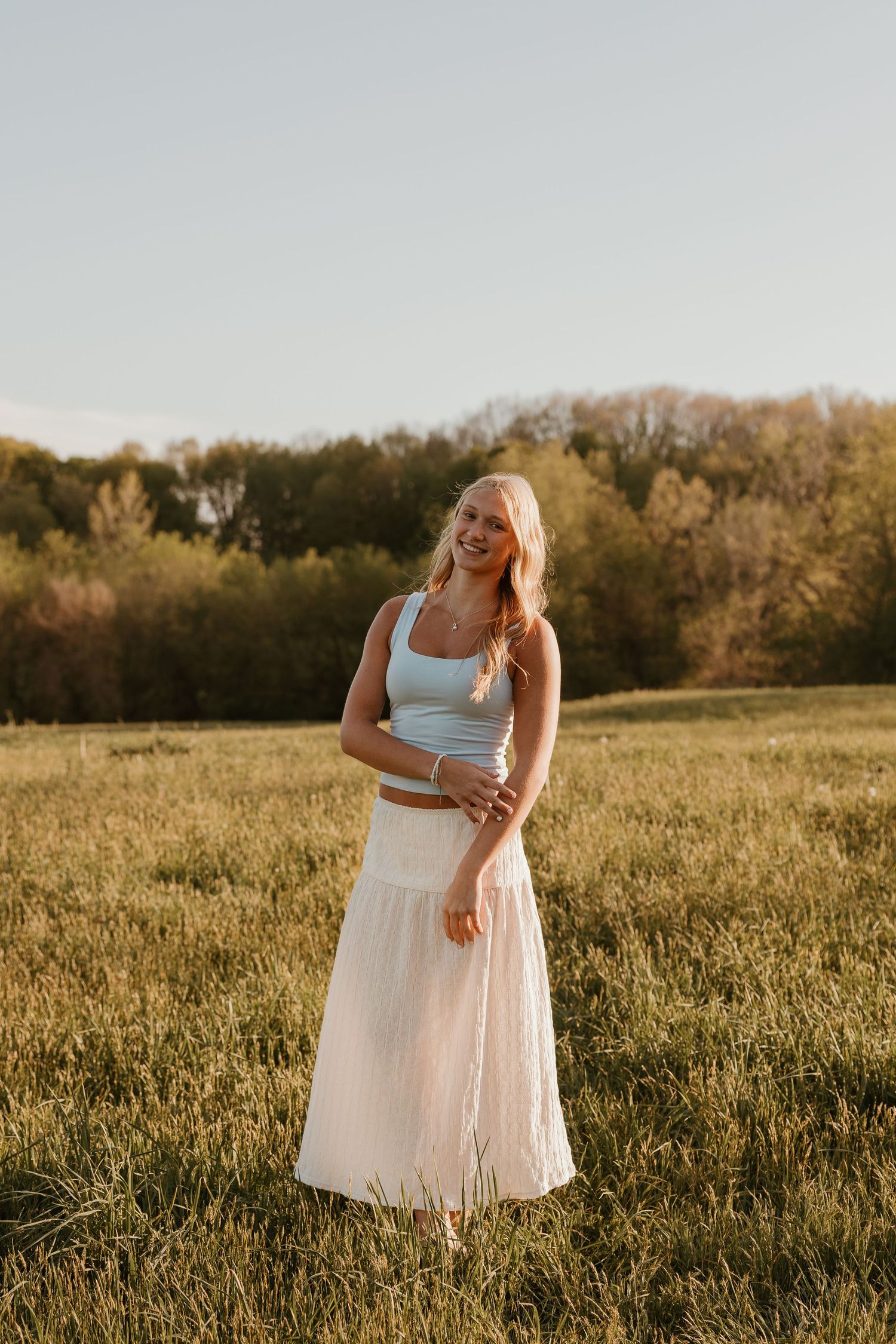 Smiling blonde woman in light blue crop top and white maxi skirt standing in a sunlit green field.
