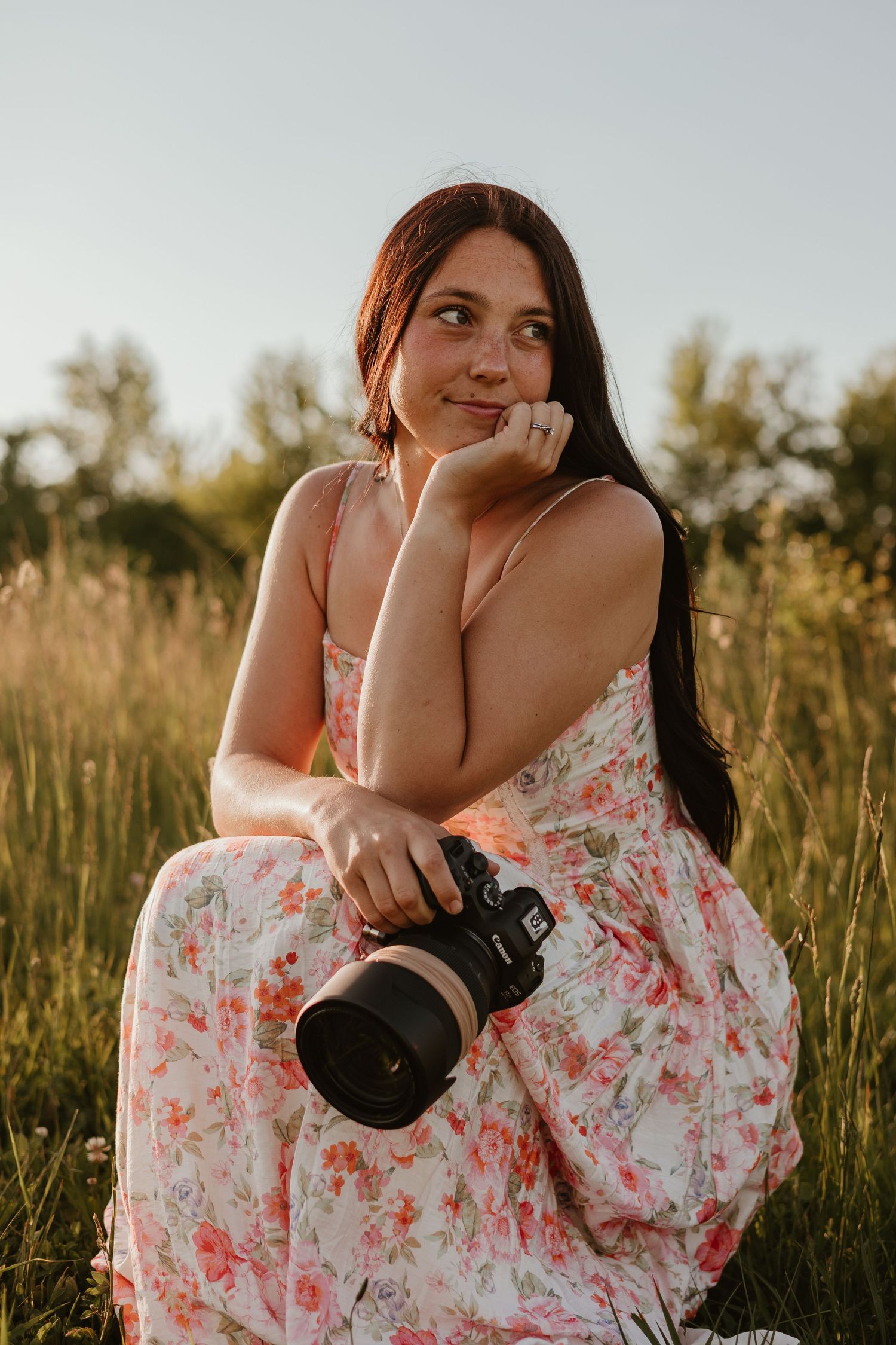 Young woman in floral dress holding a camera sits in a sunlit field, gazing thoughtfully to the side.