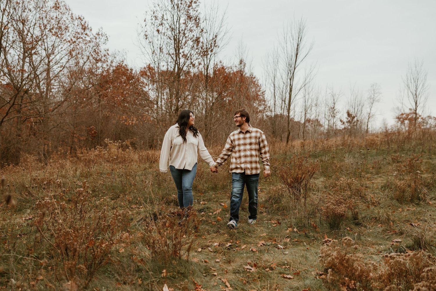 Couple holding hands walking through autumn forest trail with colorful fall foliage and bare trees.