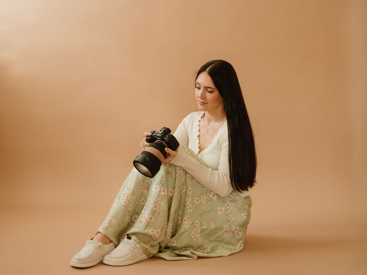 Woman in floral dress and white cardigan sits on floor holding a camera against a beige background.