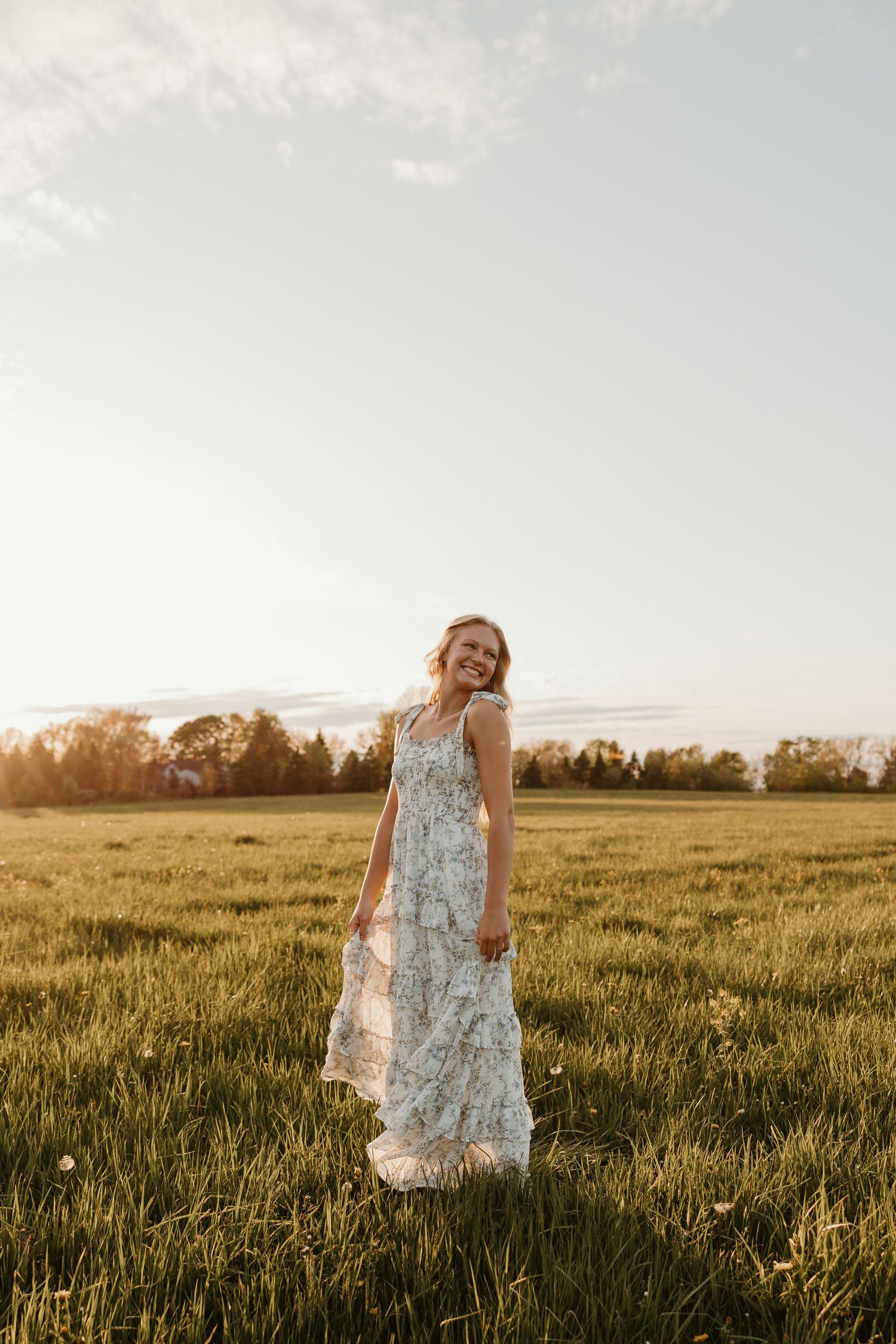Smiling blonde woman in floral maxi dress standing in green field at golden hour sunset.