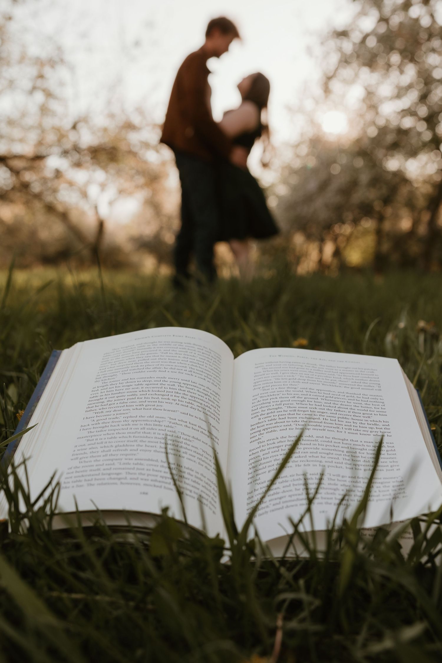 An open book rests on green grass with a blurred couple dancing romantically in a sunlit garden background.