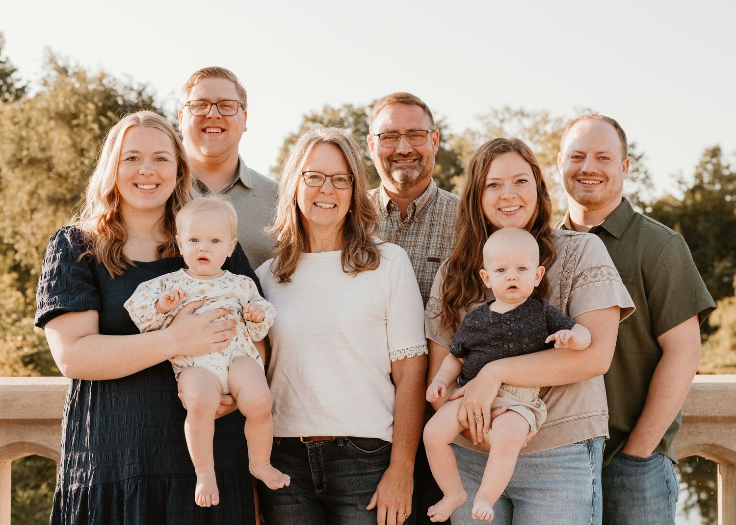 A happy family of seven poses outdoors, including two babies, smiling together on a sunny day.