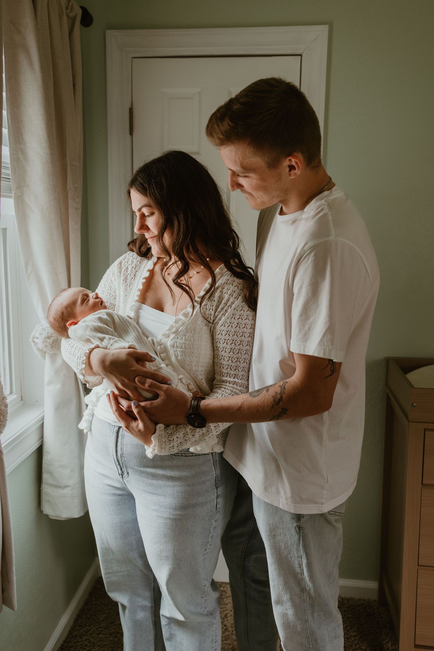 Young couple in casual attire lovingly gazing at their newborn baby by a window in a softly lit room.