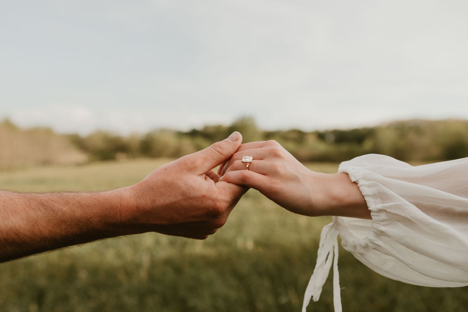 Couple holding hands outdoors, woman wearing an engagement ring, with a green field in the background.