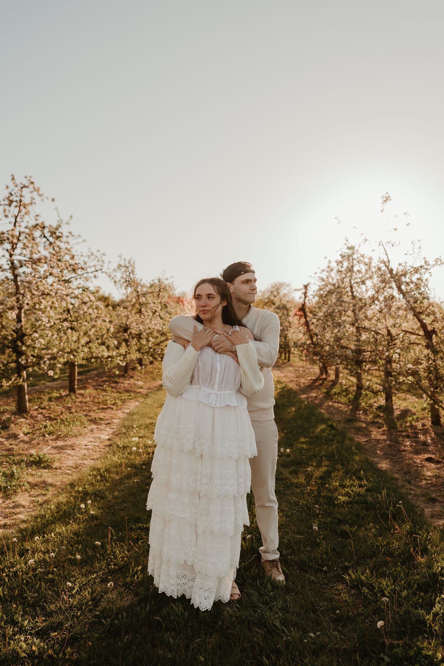 Couple embracing in a blooming orchard, bride in white lace dress, groom in tan suit, golden hour light.