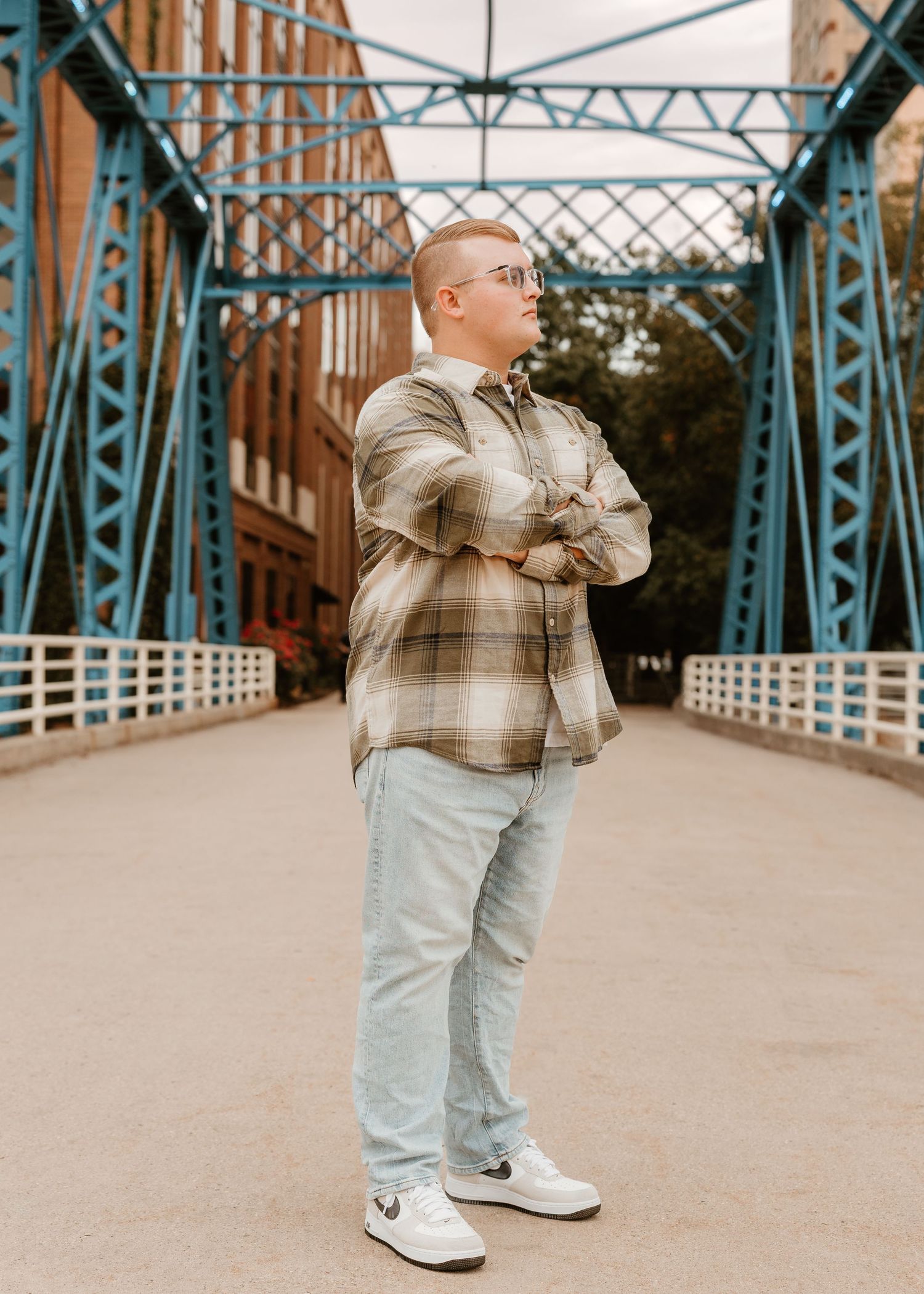 Young man in plaid shirt and light jeans stands arms crossed on a blue steel bridge, wearing white sneakers.