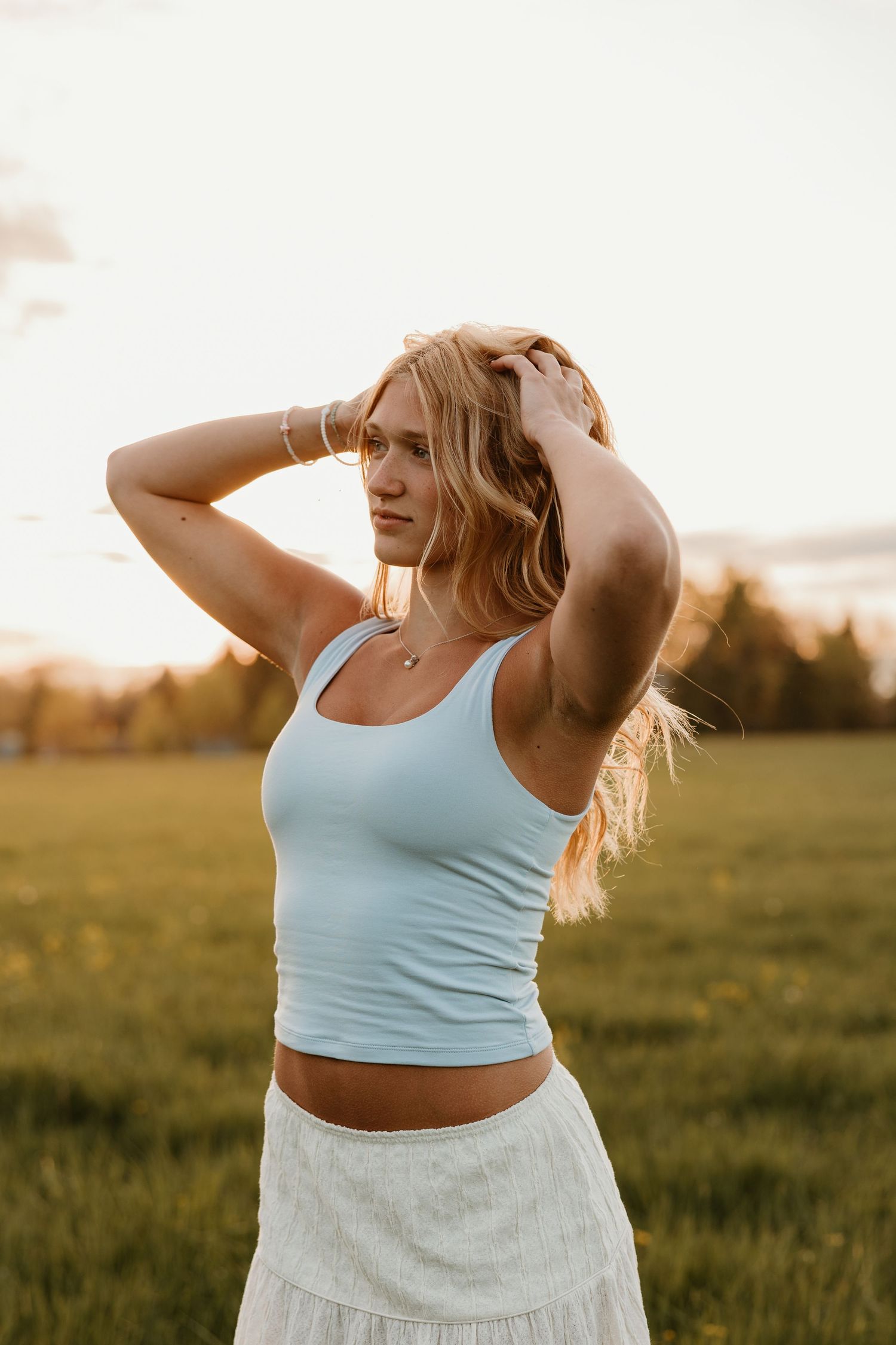 Young blonde woman in light blue crop top and white skirt poses in golden hour sunlight in a green field.