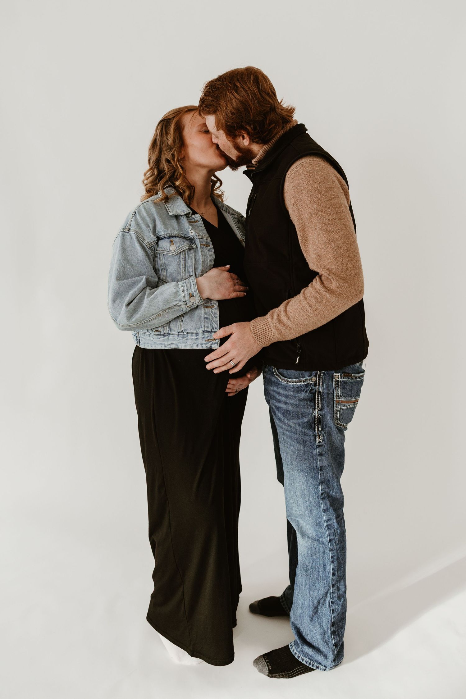 Pregnant couple sharing a kiss in a studio, woman in black dress and denim jacket, man in vest and jeans.