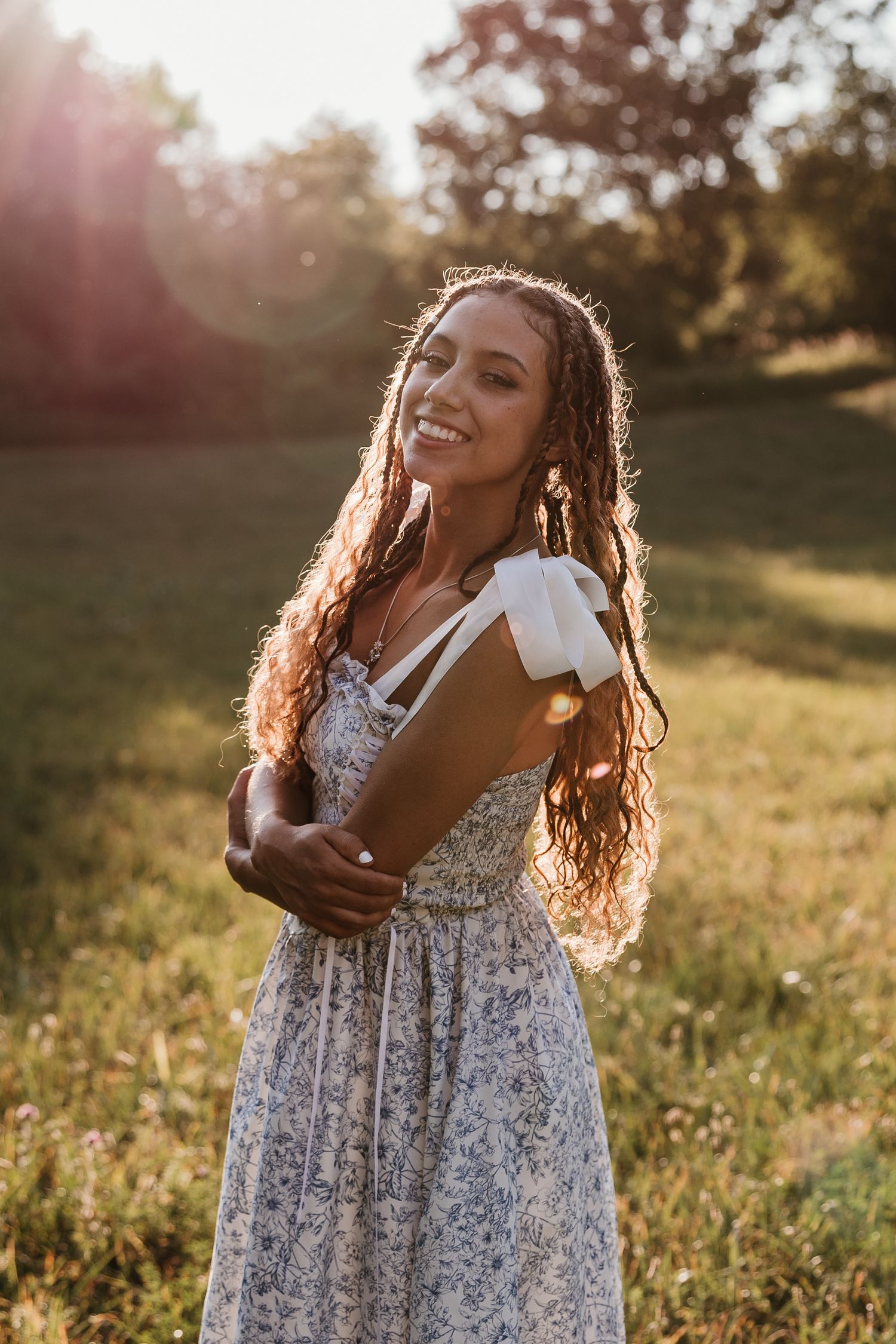 Smiling young woman in floral sundress holding books outdoors in golden sunset light with trees in background.