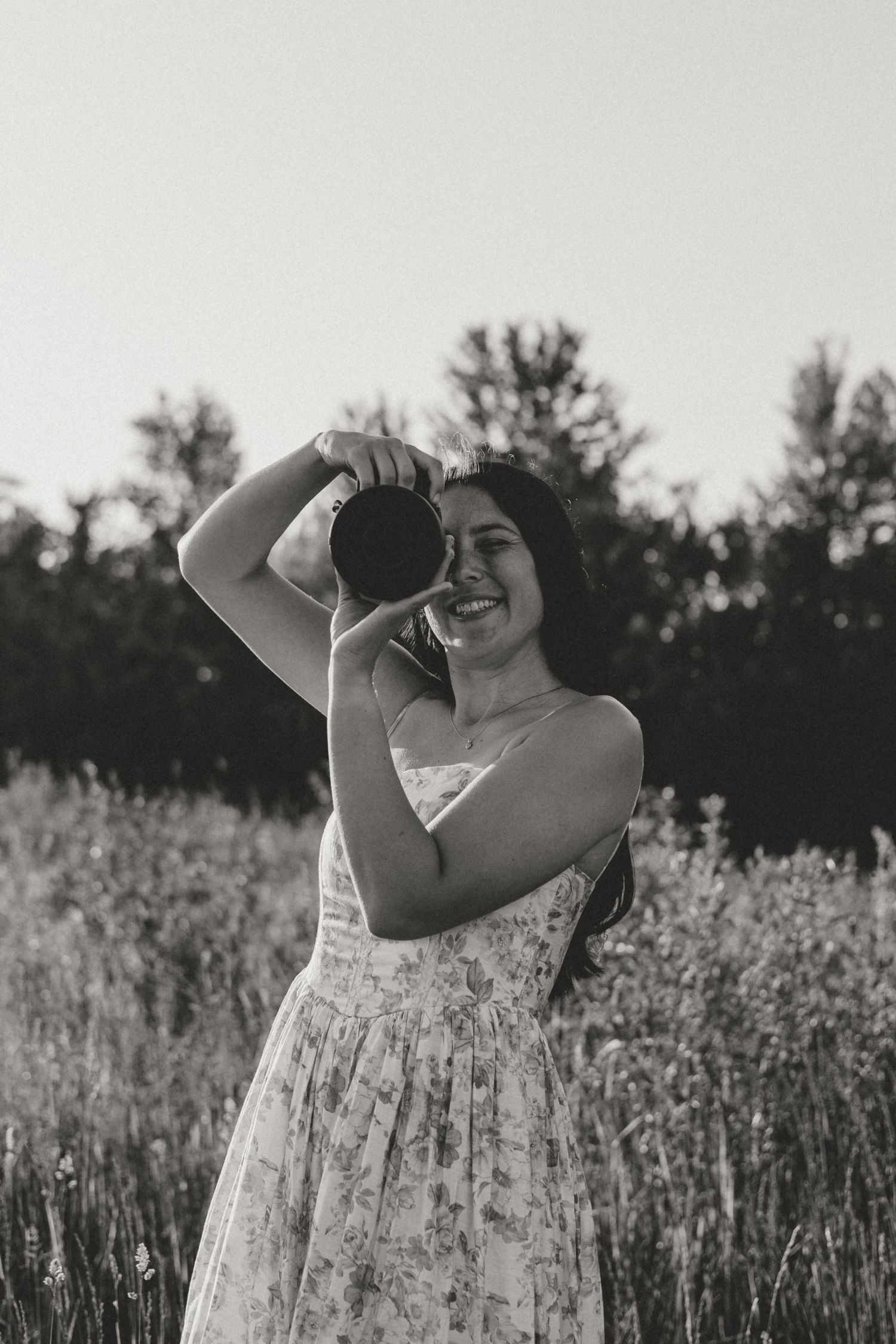 Black and white photo of a smiling woman in a floral dress holding a camera in a wildflower field.