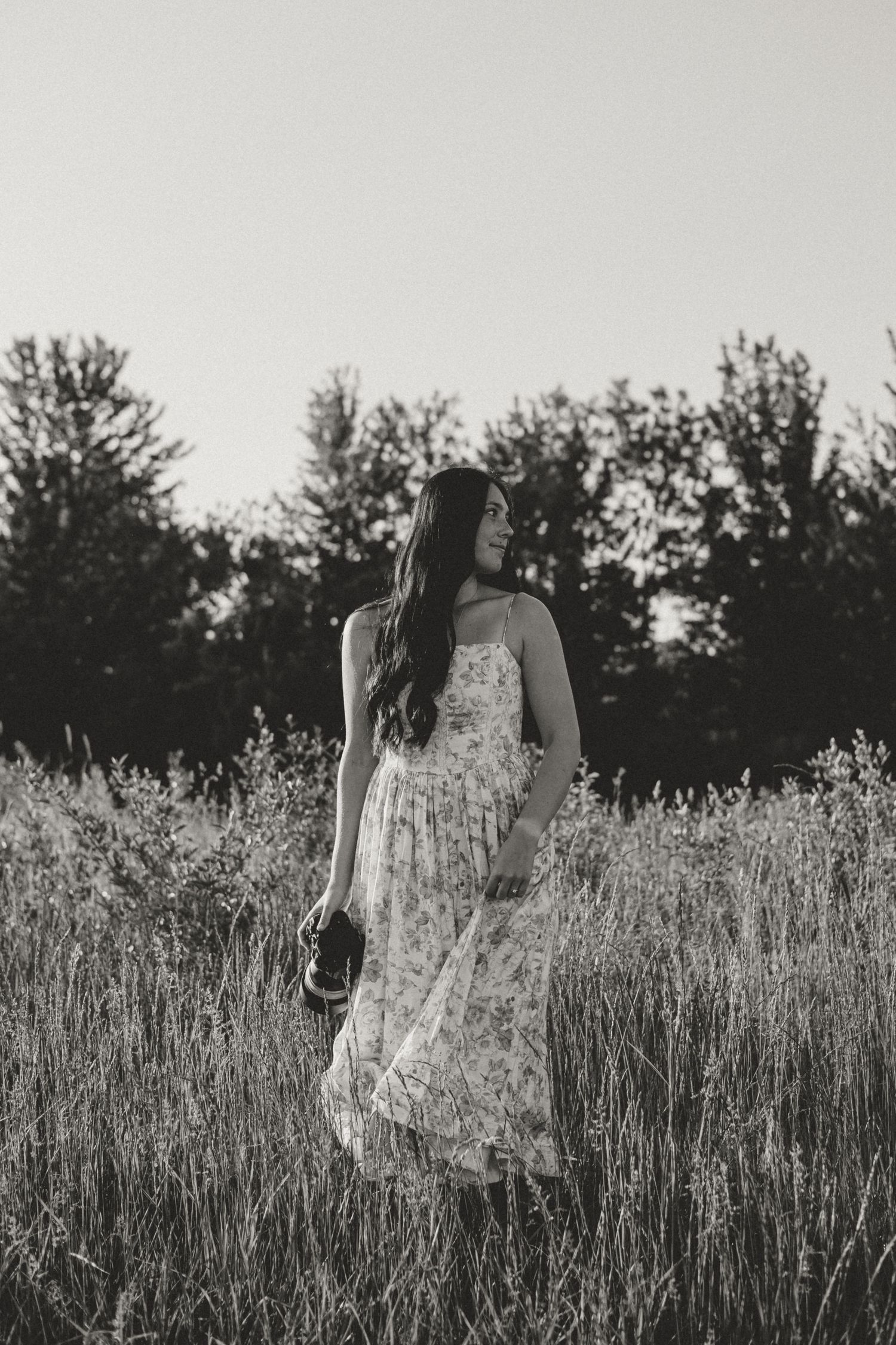 Black and white photo of a young woman in a floral dress standing in a tall grass field with trees in the background.