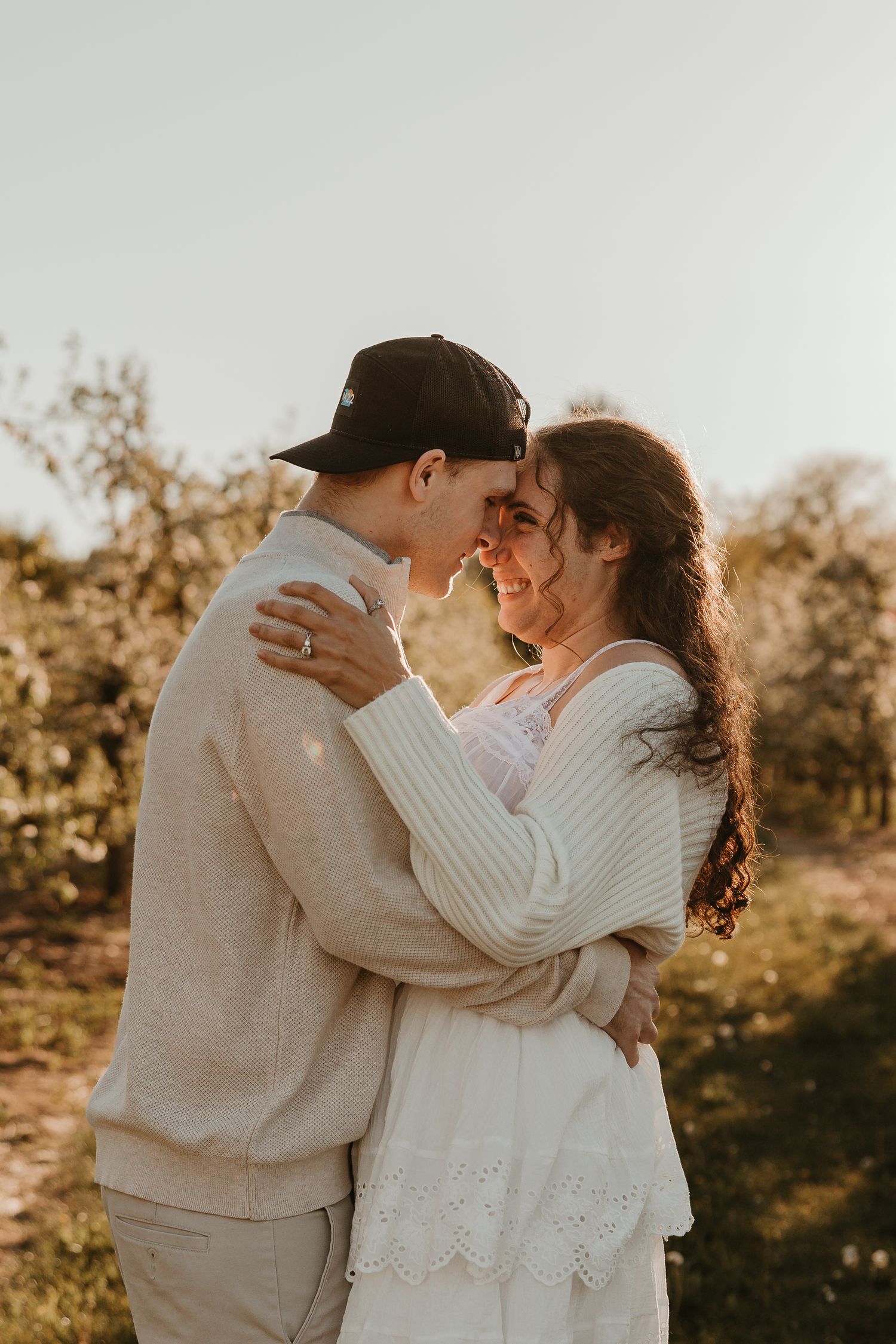 Couple embracing outdoors at golden hour, wearing neutral tones, sharing a romantic moment surrounded by blooming trees.