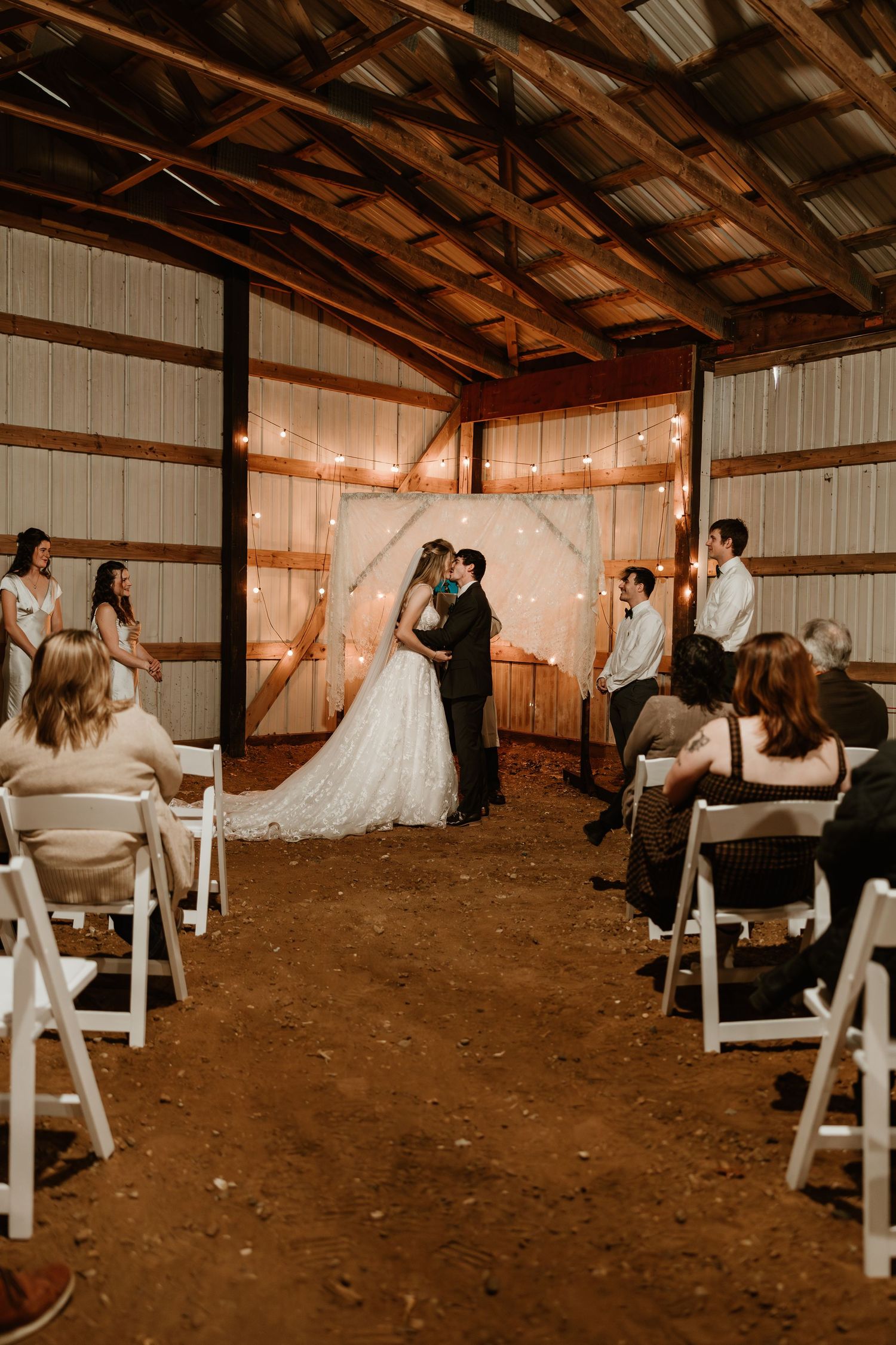 Rustic barn wedding ceremony with bride and groom kissing at altar adorned with string lights and floral arch.