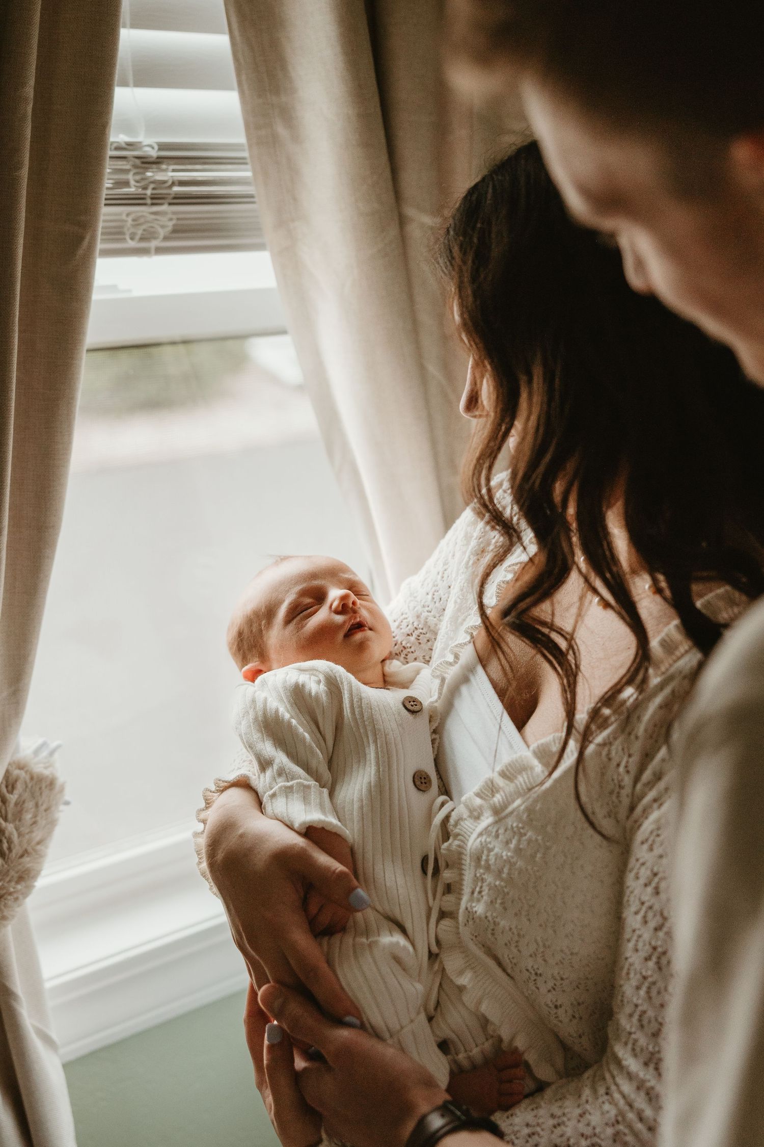 Mother in white lace dress tenderly holding newborn baby by a sunlit window.