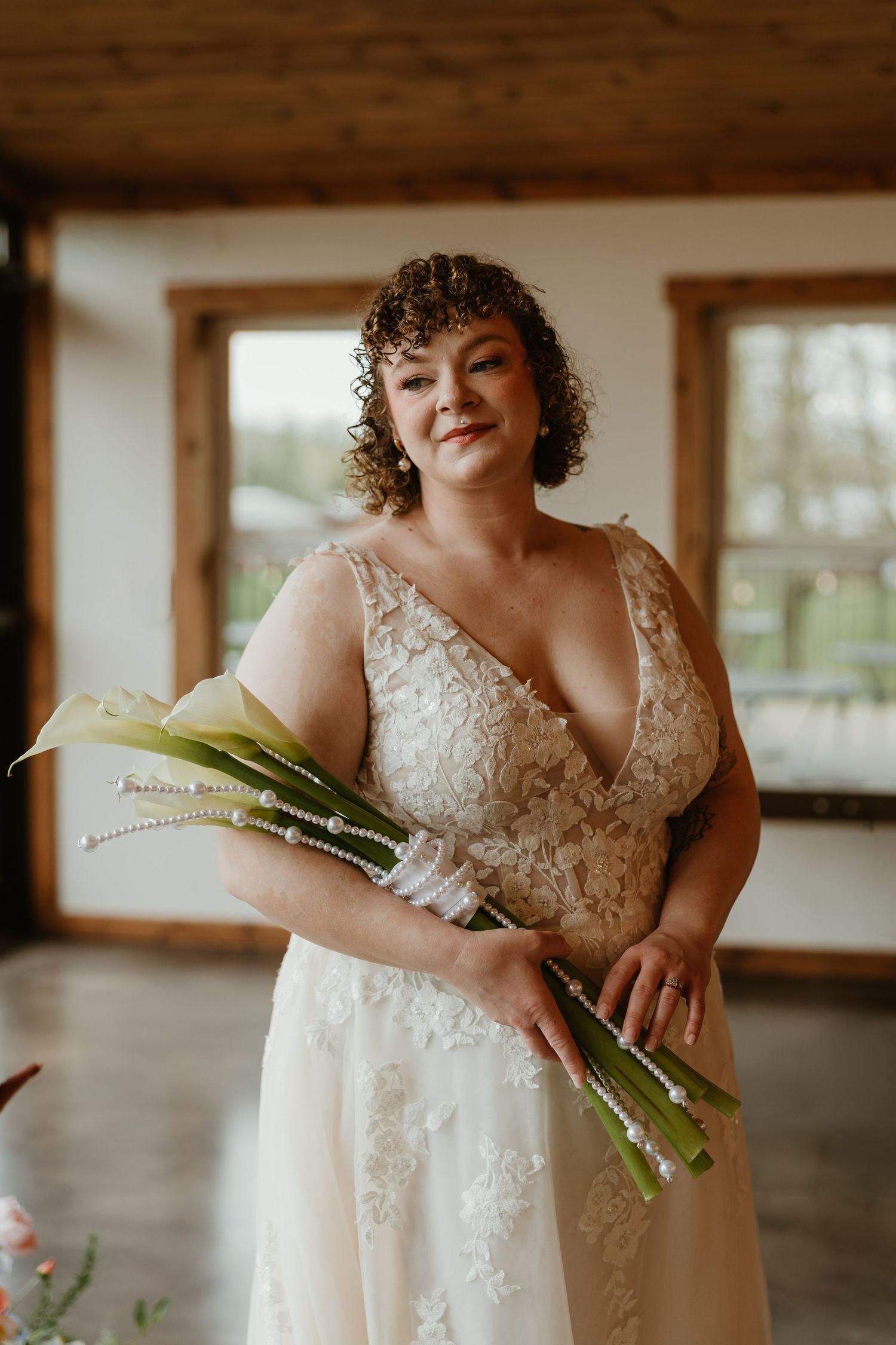 Bride in lace wedding gown holding calla lily bouquet with pearl wrap, smiling in rustic barn venue.