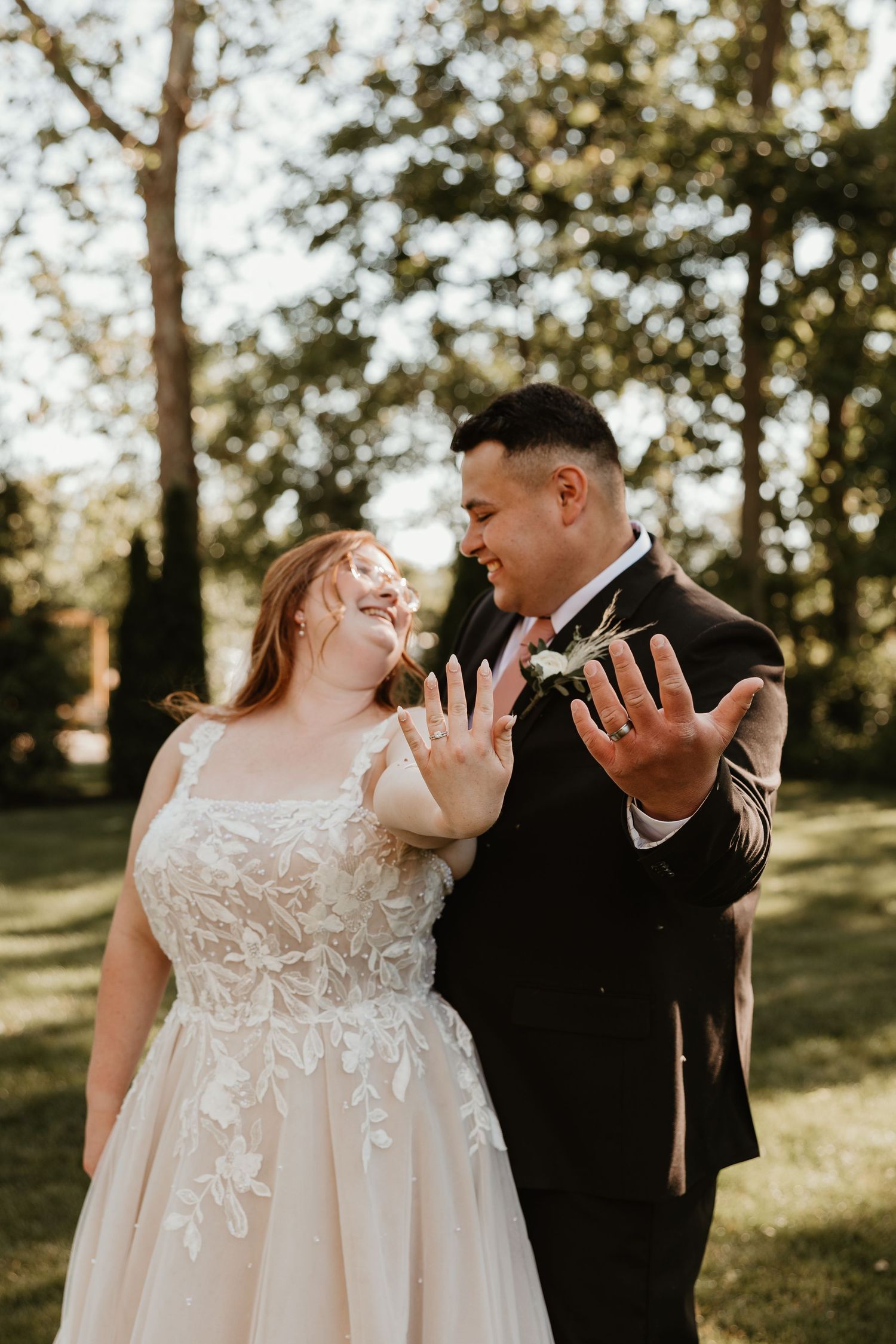Newlywed couple smiling and showing off wedding rings outdoors, bride in lace gown and groom in black tuxedo.